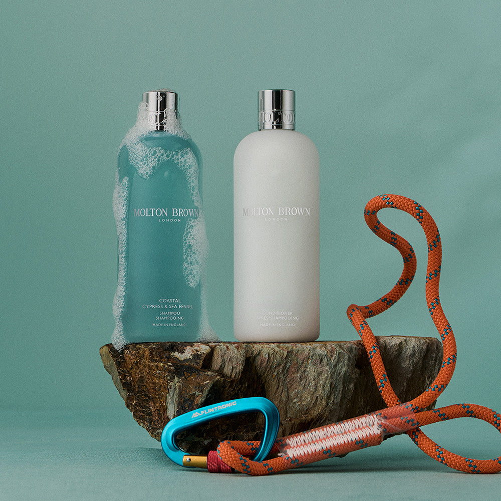 Two Molton Brown Cyprus bottles sit on a rough stone slab—one white Coastal Cypress & Sea Fennel Conditioner 300ml, the other blue-green. An orange climbing rope and blue carabiner lie in front, set against a green background.
