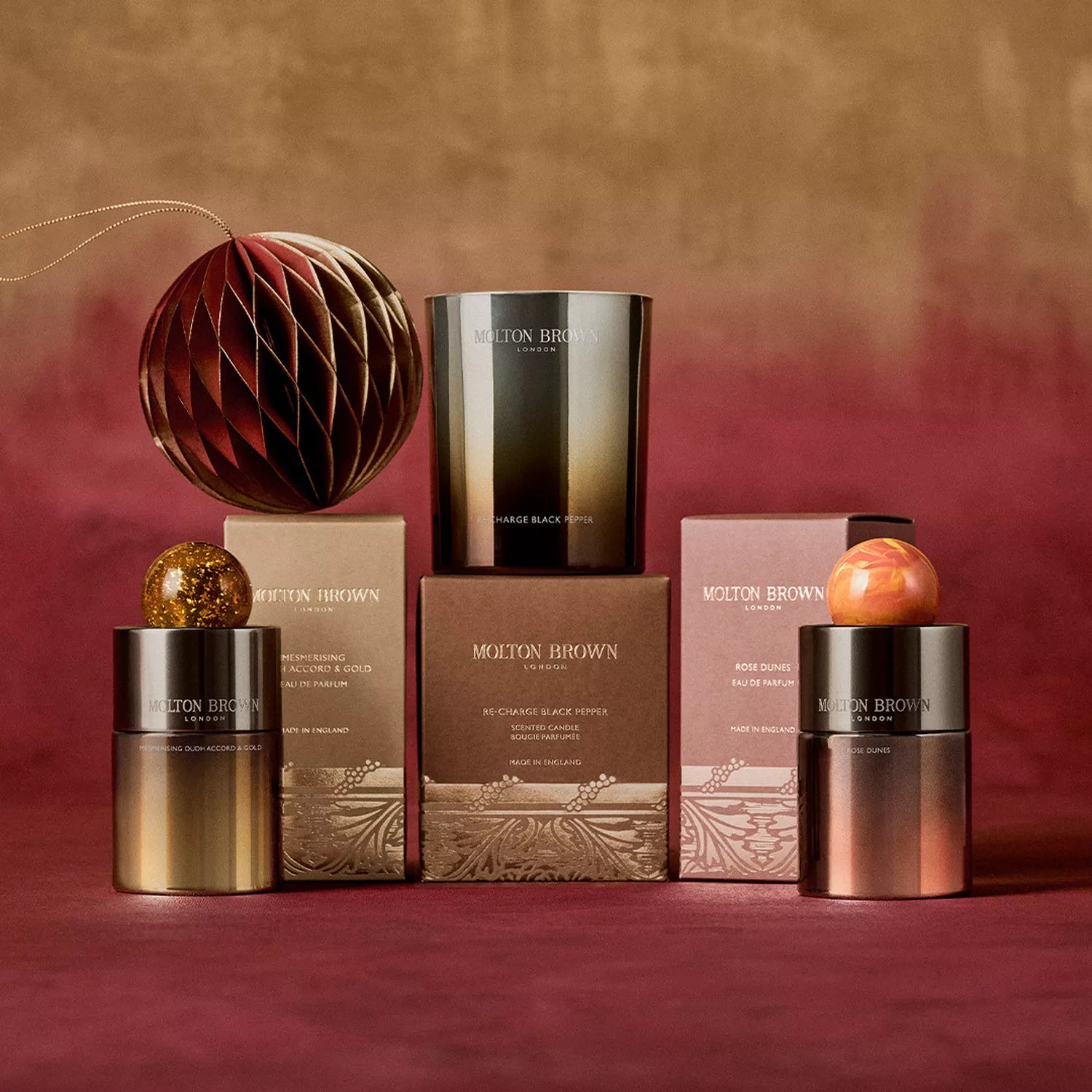 A Limited Edition Re-charge Black Pepper Signature Candle 190g by MoltonBrown Cyprus is displayed with festive ornaments on a red and gold surface. The burgundy-to-gold background creates a warm, spiced atmosphere ideal for the season.