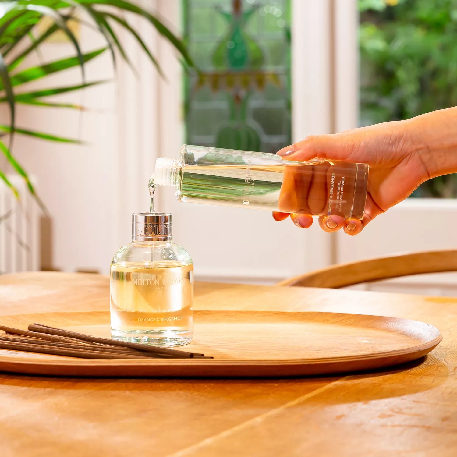 A hand gracefully pours Sample 3 from MoltonBrown Cyprus out of a clear bottle into a round glass container on a wooden tray. Nearby, MoltonBrown Cyprus aroma reeds stand ready to infuse the air. A plant and window provide a serene backdrop.