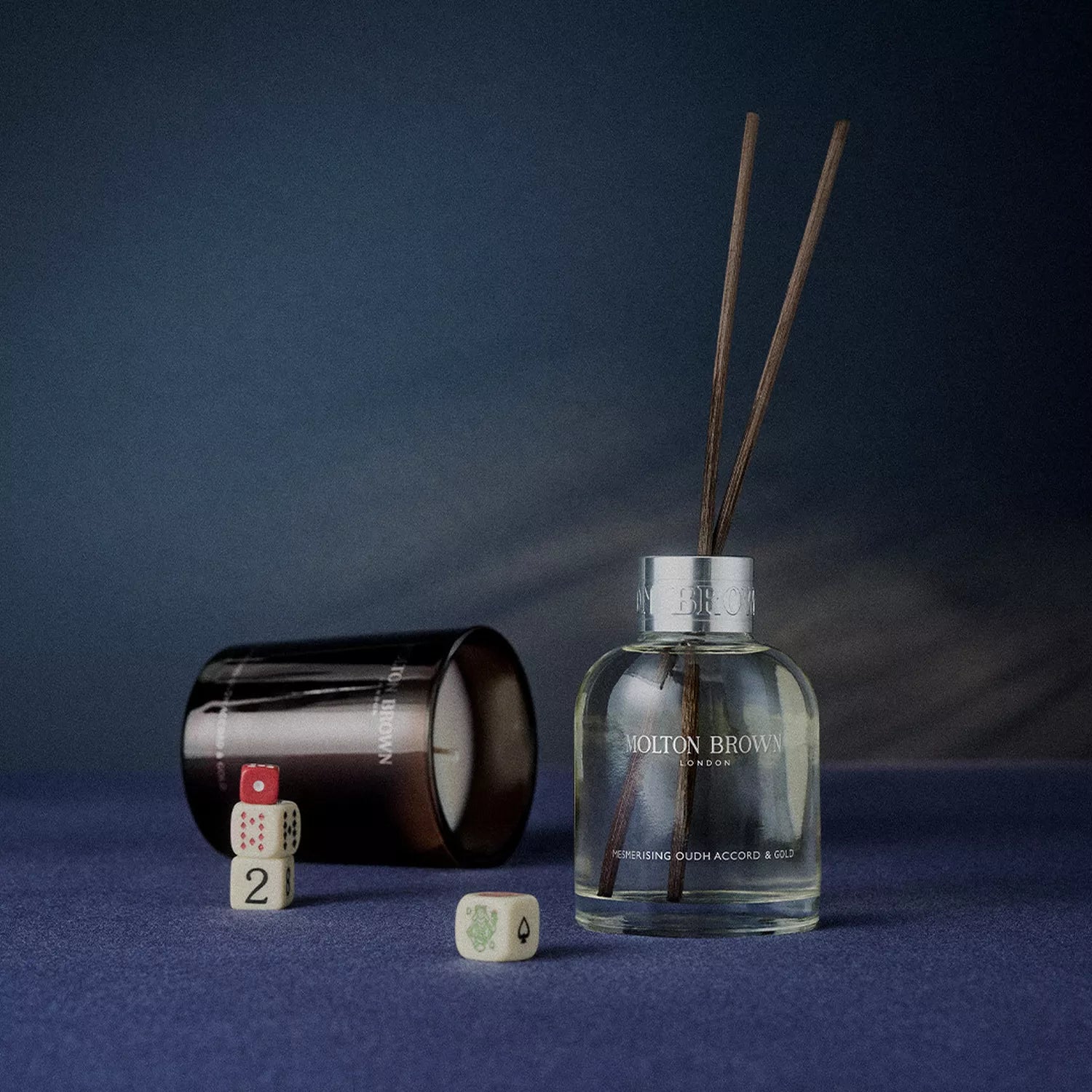 A MoltonBrown Cyprus Mesmerising Oudh Accord & Gold Aroma Reeds 150ml diffuser with three sticks sits on a blue surface, radiating luxury. Beside it, a dark glass container rests on its side with three game dice scattered across the gradient blue background.