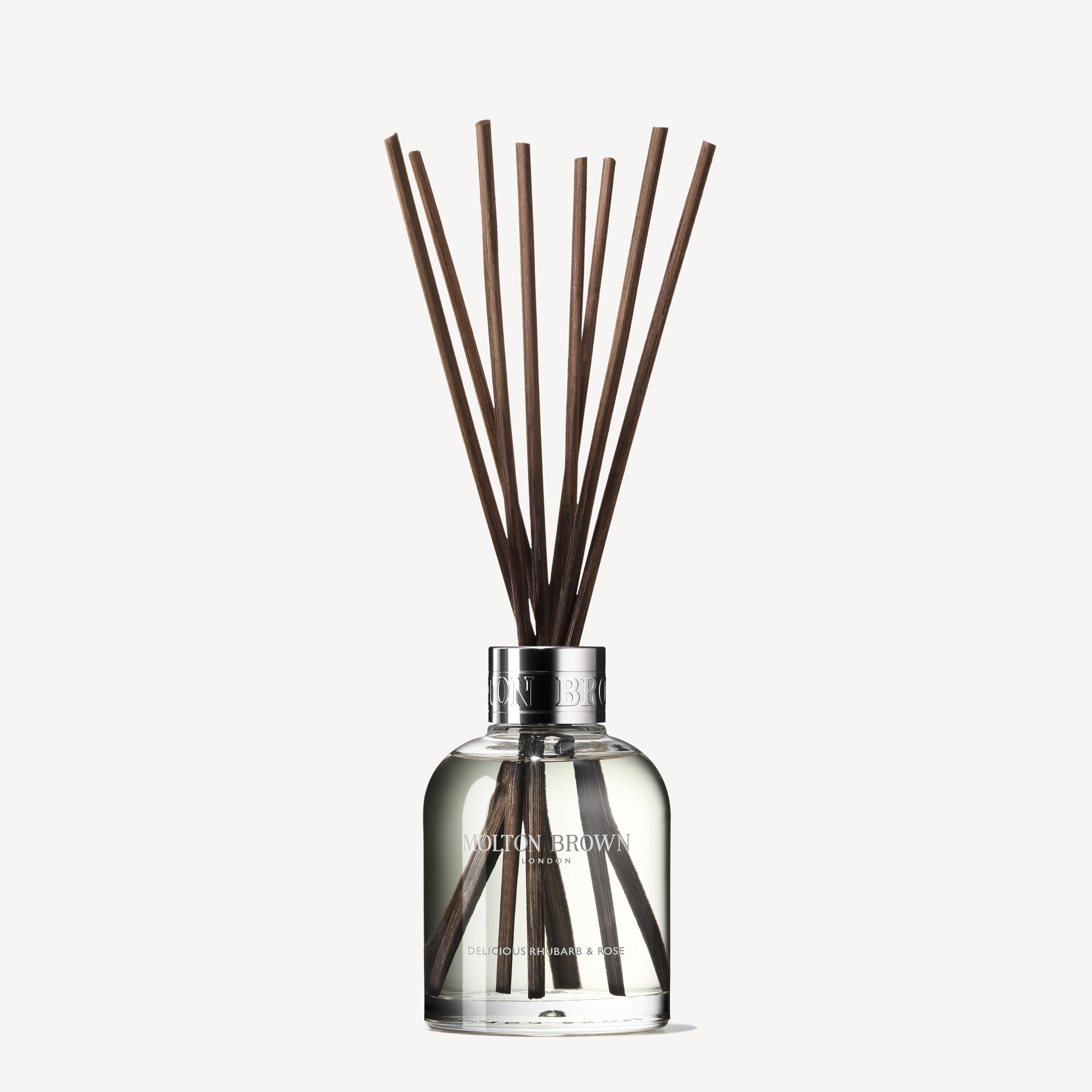 The Delicious Rhubarb & Rose Aroma Reeds 150ml from MoltonBrown Cyprus comes in a clear glass bottle, showcasing several long, dark brown reeds that extend upward. The visible liquid emits enticing exotic rhubarb and rose scents. A silver cap adorned with the brand name completes this sophisticated accessory, promising a luxurious aroma.