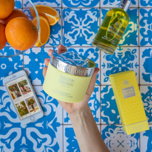 A hand holds the Orange & Bergamot Radiant Body Polisher 275g from MoltonBrown Cyprus, a yellow luxury body cream with citrus notes, above blue and white patterned tiles. Nearby, a matching yellow box, a bottle of perfume, and fresh oranges sit in a bowl to the side.