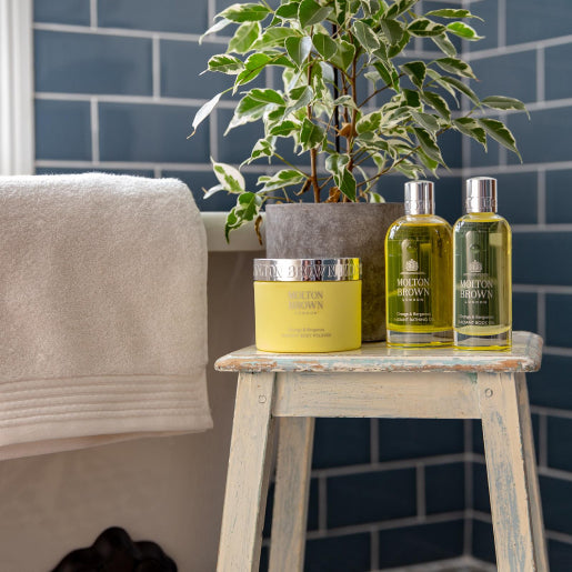 A cozy bathroom scene features a wooden stool displaying a jar of MoltonBrown Cyprus' Orange & Bergamot Radiant Body Polisher and two bottles of Molton Brown shower gel. A potted plant adds greenery, while soft towels hang nearby against a backdrop of dark blue subway tiles, inviting relaxation and rejuvenation with citrus-infused luxury.