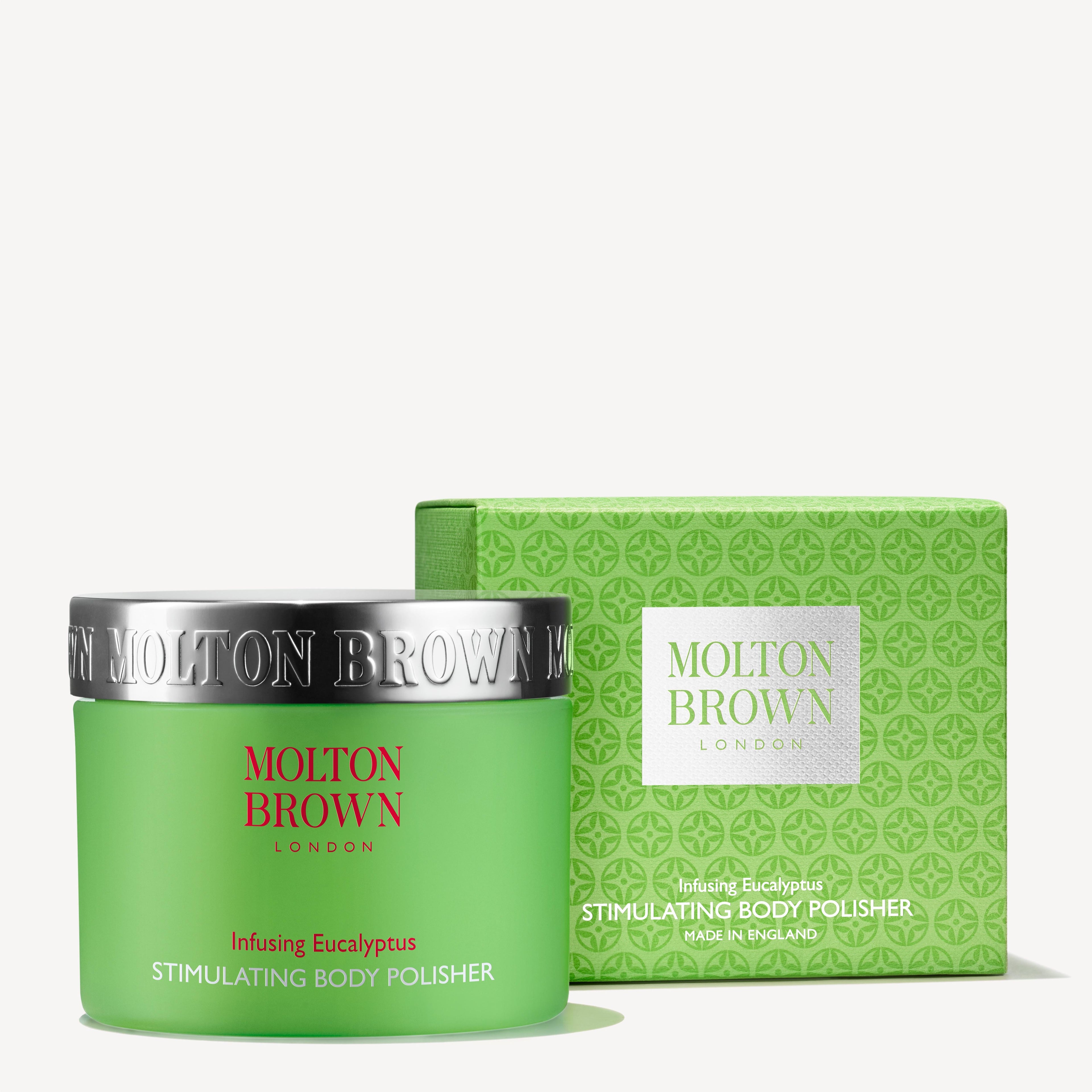 A green container of MoltonBrown Cyprus Infusing Eucalyptus Stimulating Body Polisher 275g, an exfoliating sugar body scrub, is positioned next to its coordinating green box. Both display silver and red text that detail the product's ability to purify and smooth skin.