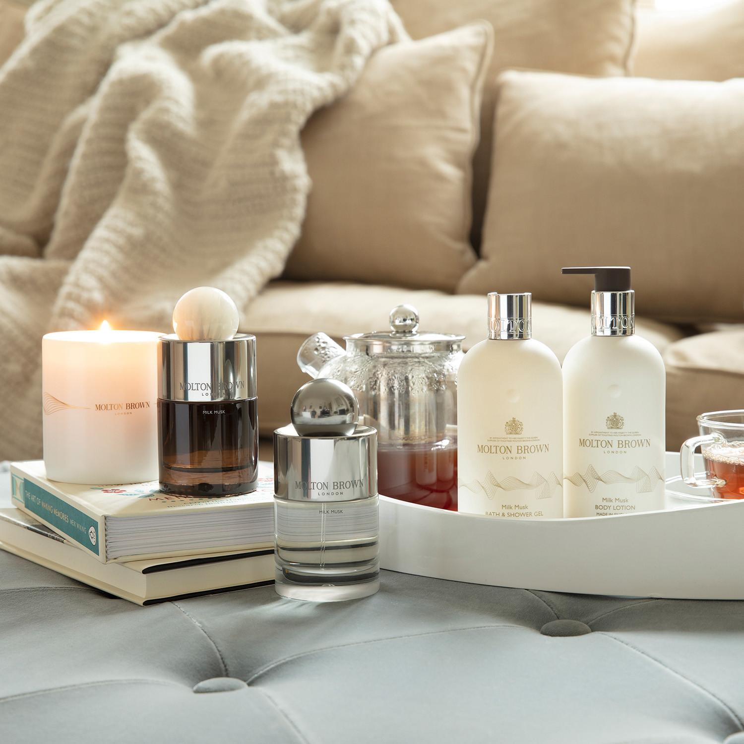 A cozy setting with MoltonBrown Cyprus products, featuring a Milk Musk Bath & Shower Gel 300ml along with lotions and a candle on a tray. Nearby, tea is served in a glass pot and cup placed on a plush ottoman surrounded by a stack of books, while soft, beige cushions create an inviting atmosphere in the background.