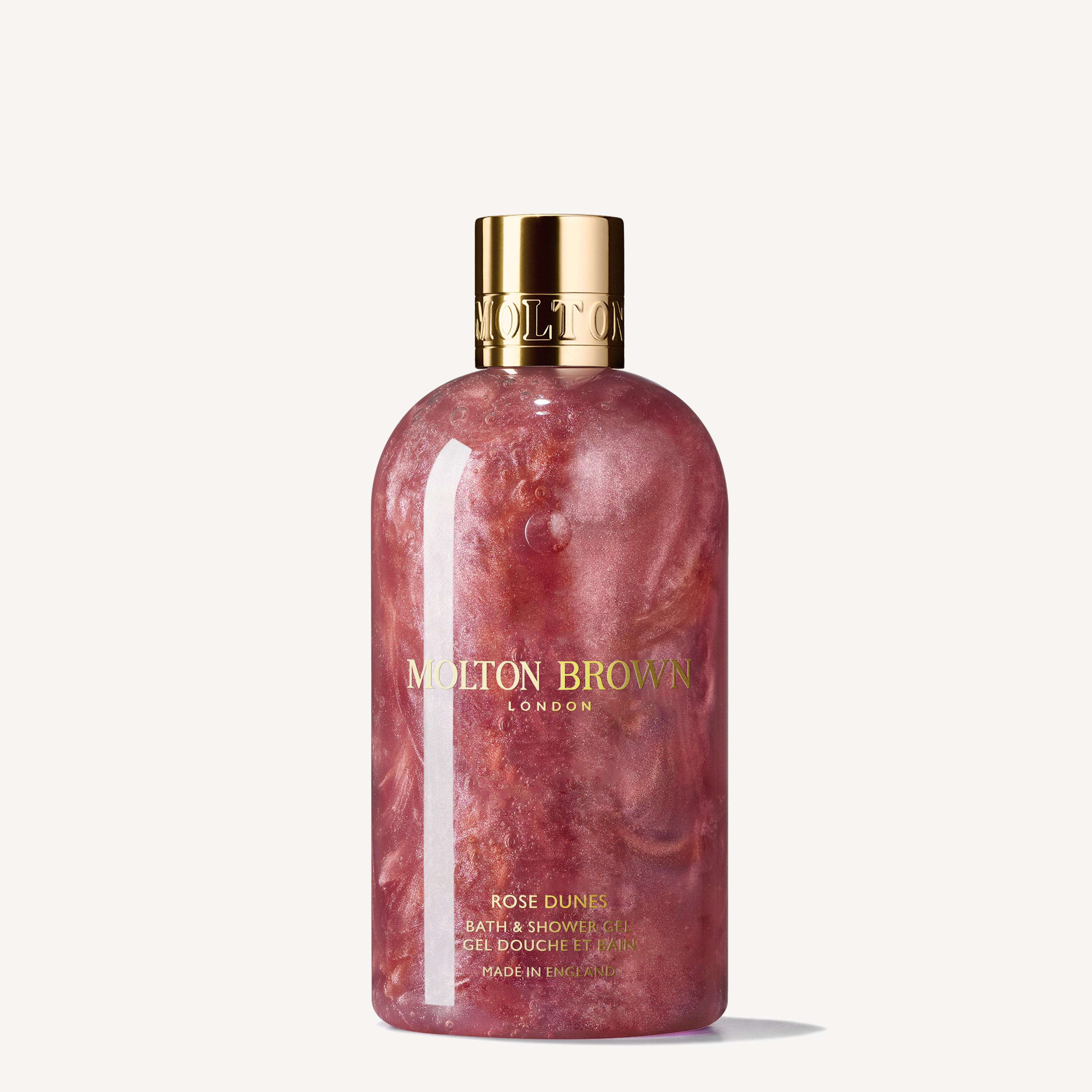 The Rose Dunes Bath & Shower Gel 300ml from MoltonBrown Cyprus features a pink, marbled liquid infused with patchouli Sulawesi, and comes with a gold cap and label adorned with elegant branding and text.