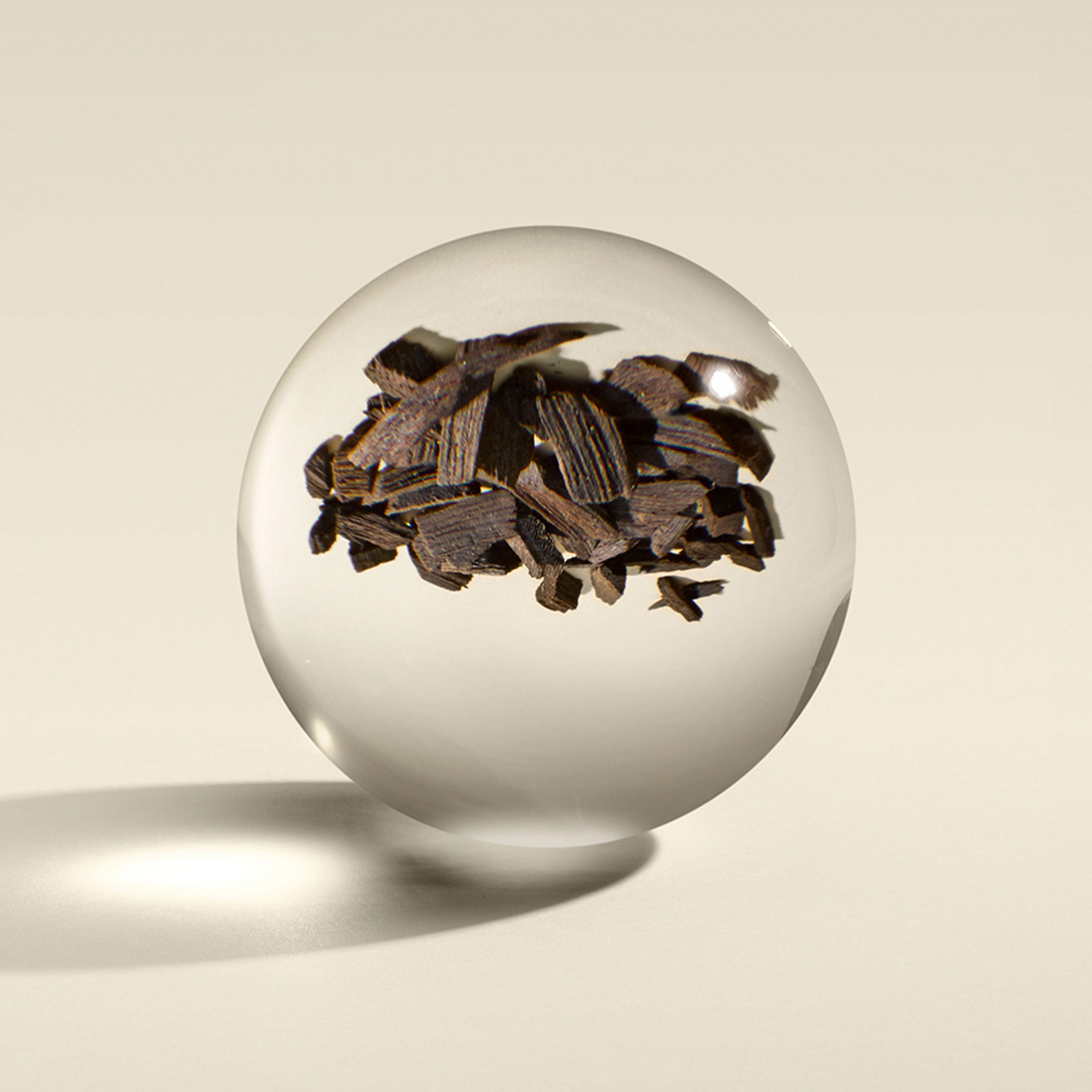 A transparent sphere encloses dark wood chips, casting a shadow on a light beige surface. Suspended at the center, the chips seem to float mysteriously, reminiscent of 23.5-carat golden flakes shimmering in Molton Brown Cyprus's Mesmerising Oudh Accord & Gold Fine Liquid Hand Wash.