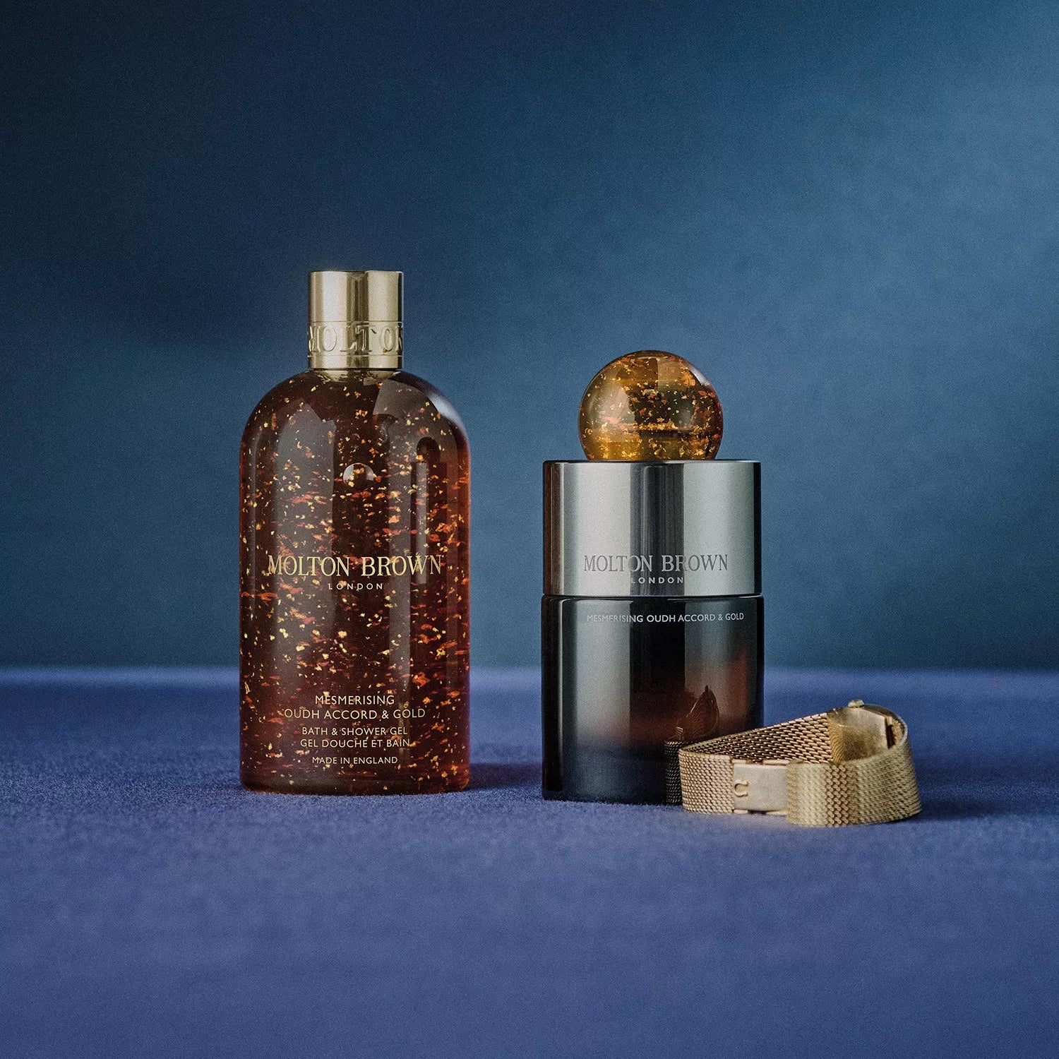 A MoltonBrown Cyprus Mesmerising Oudh Accord & Gold Eau de Parfum 100ml in a collectible glass flacon with gold cap sits beside a bath and shower gel bottle and gold mesh watch on a blue surface, against a blue background.