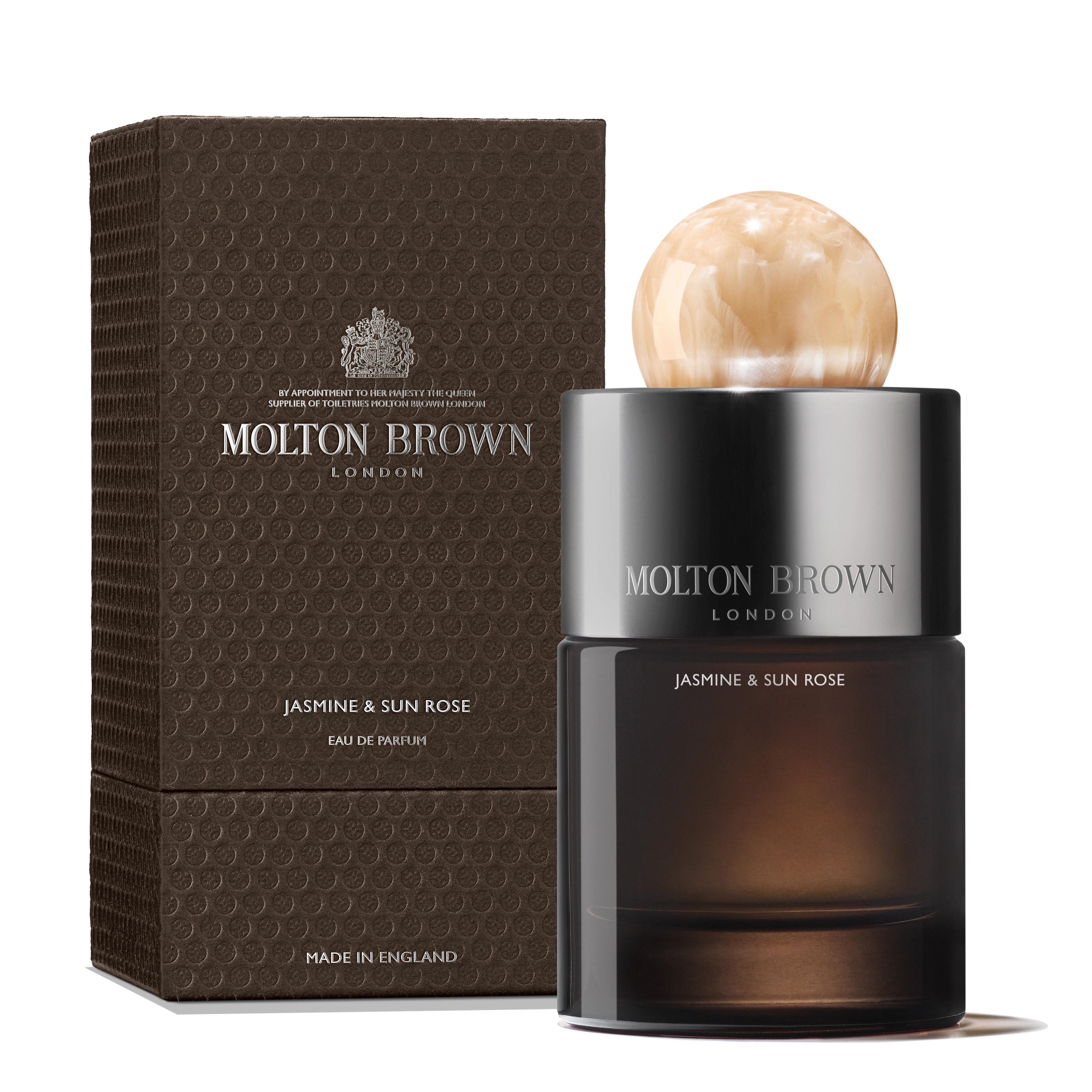 The MoltonBrown Cyprus Jasmine & Sun Rose Eau de Parfum 100ml, featuring a marble-like round cap, elegantly rests next to its textured brown packaging. The exquisite gold and white text enhances the sophistication, making it an ideal accessory for your eveningwear ensemble.