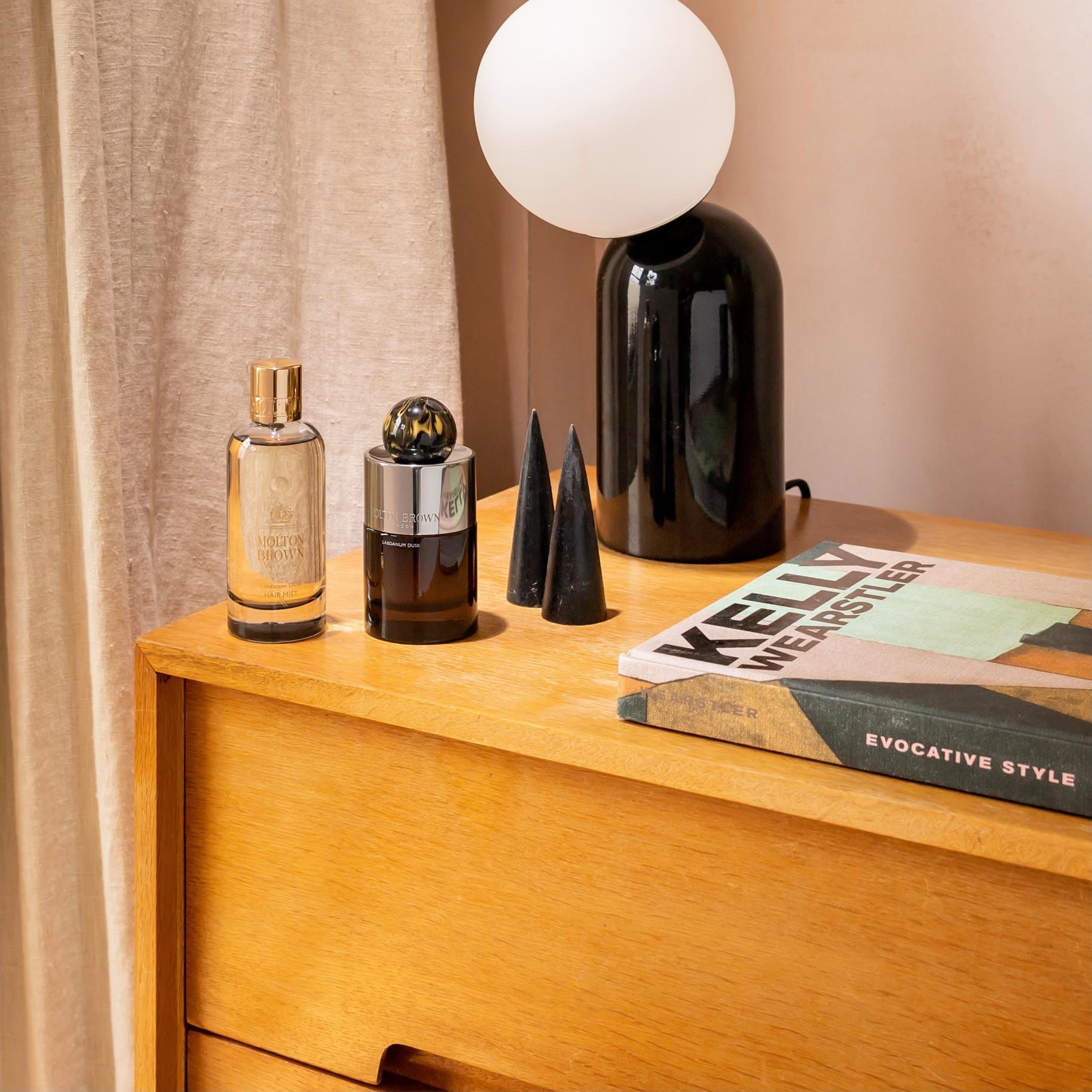 A wooden dresser holds two bottles of Labdanum Dusk Eau de Parfum by MoltonBrown Cyprus, a small black and white lamp, two black cone-shaped objects, and two books titled Kelly Wearstler and Evocative Style. An amber woody aroma subtly fills the air as a beige curtain gently frames the background.