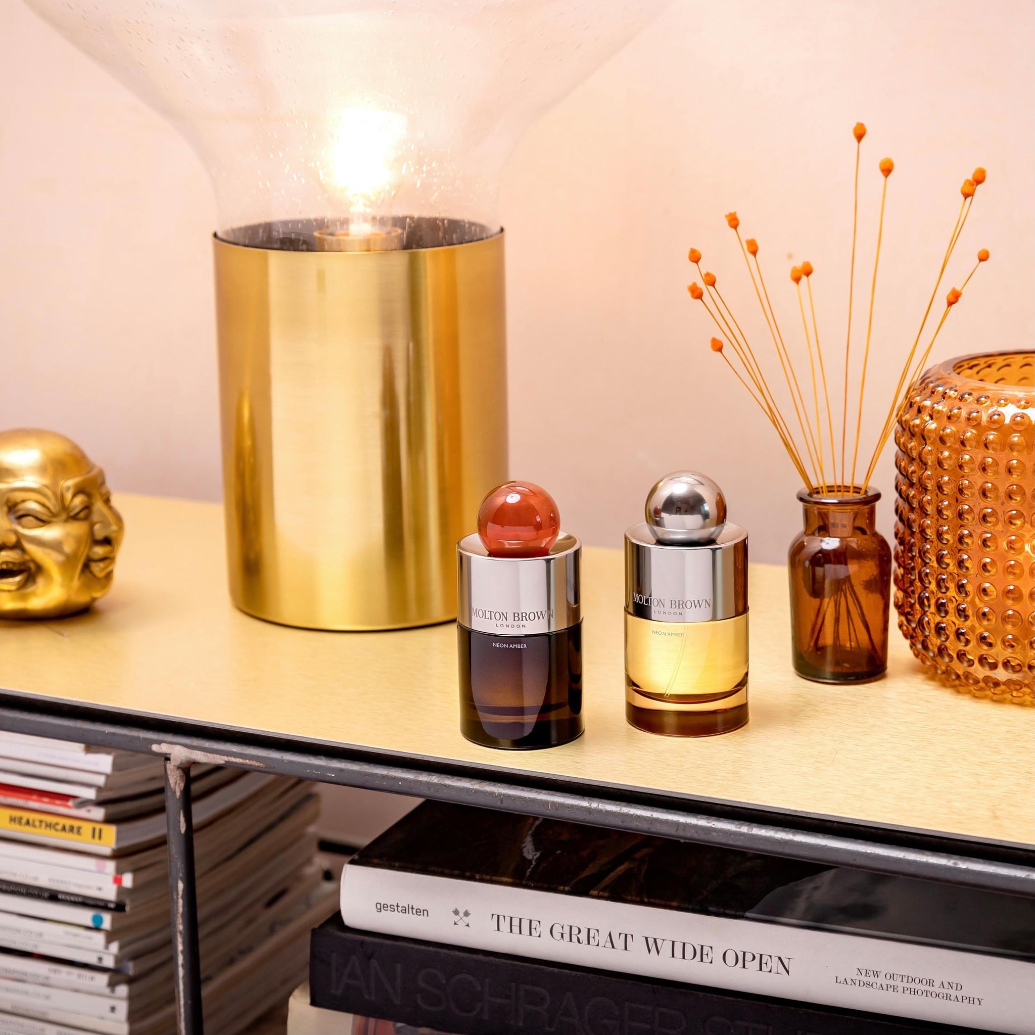 A decorative shelf showcasing a lamp with a glass shade and gold base, two bottles of MoltonBrown Cyprus products, including their Neon Amber Eau de Parfum, an orange glass vase with stems, a small gold Buddha statue, and a stack of books that includes The Great Wide Open.