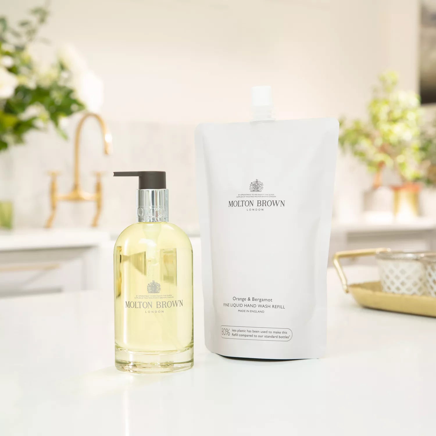 A Molton Brown Cyprus Orange & Bergamot Fine Liquid Hand Wash Glass Bottle 200ml and a refill pouch are on the kitchen counter. The transparent glass bottle with a pump complements the branded pouch, while greenery and decor enhance the sustainable scene.