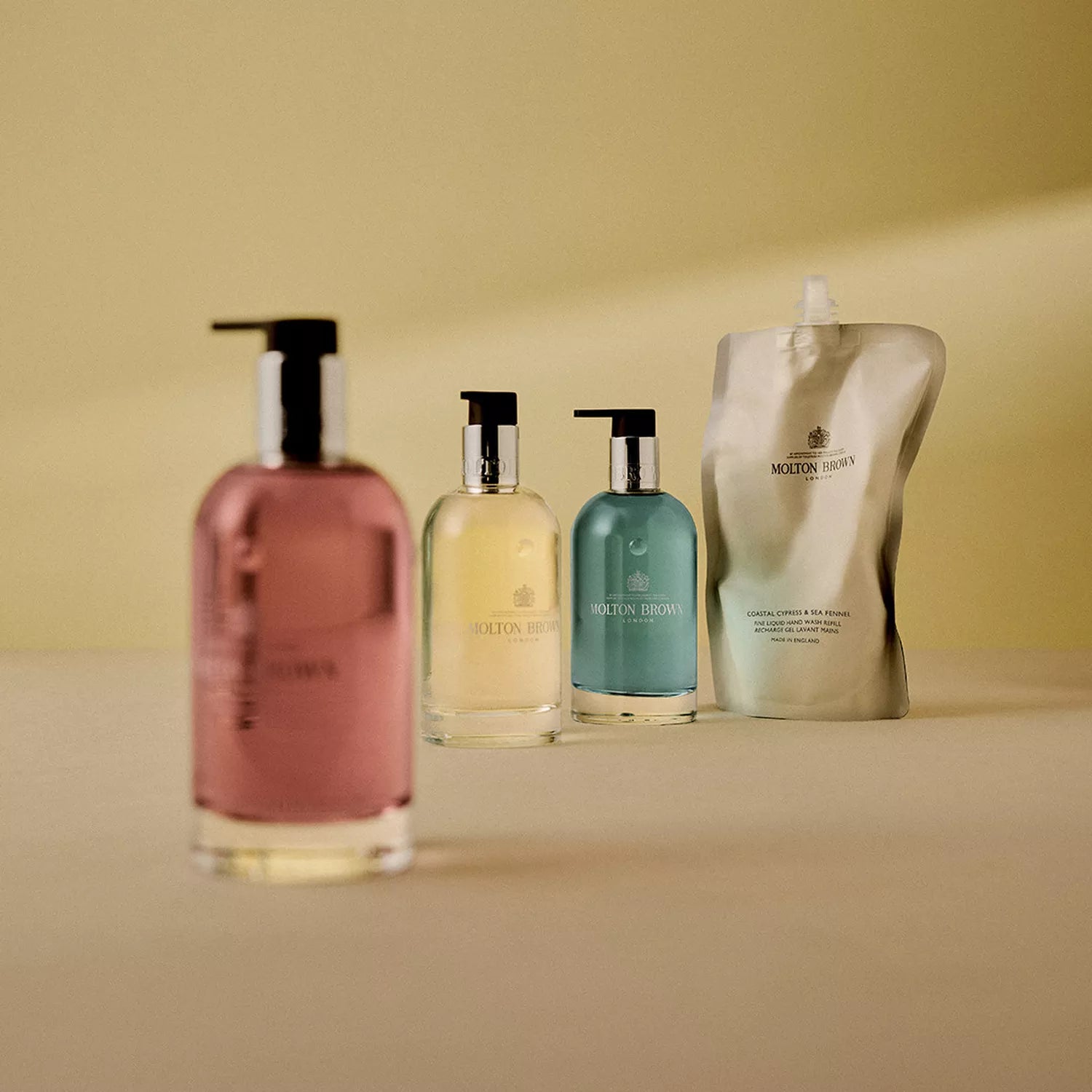 Four Molton Brown Cyprus products are aligned on a beige surface: three recyclable bottles with pumps contain pink, yellow, and Coastal Cypress & Sea Fennel Fine Liquid Hand Wash in blue. A refill pouch stands at the end against an inviting soft yellow backdrop.