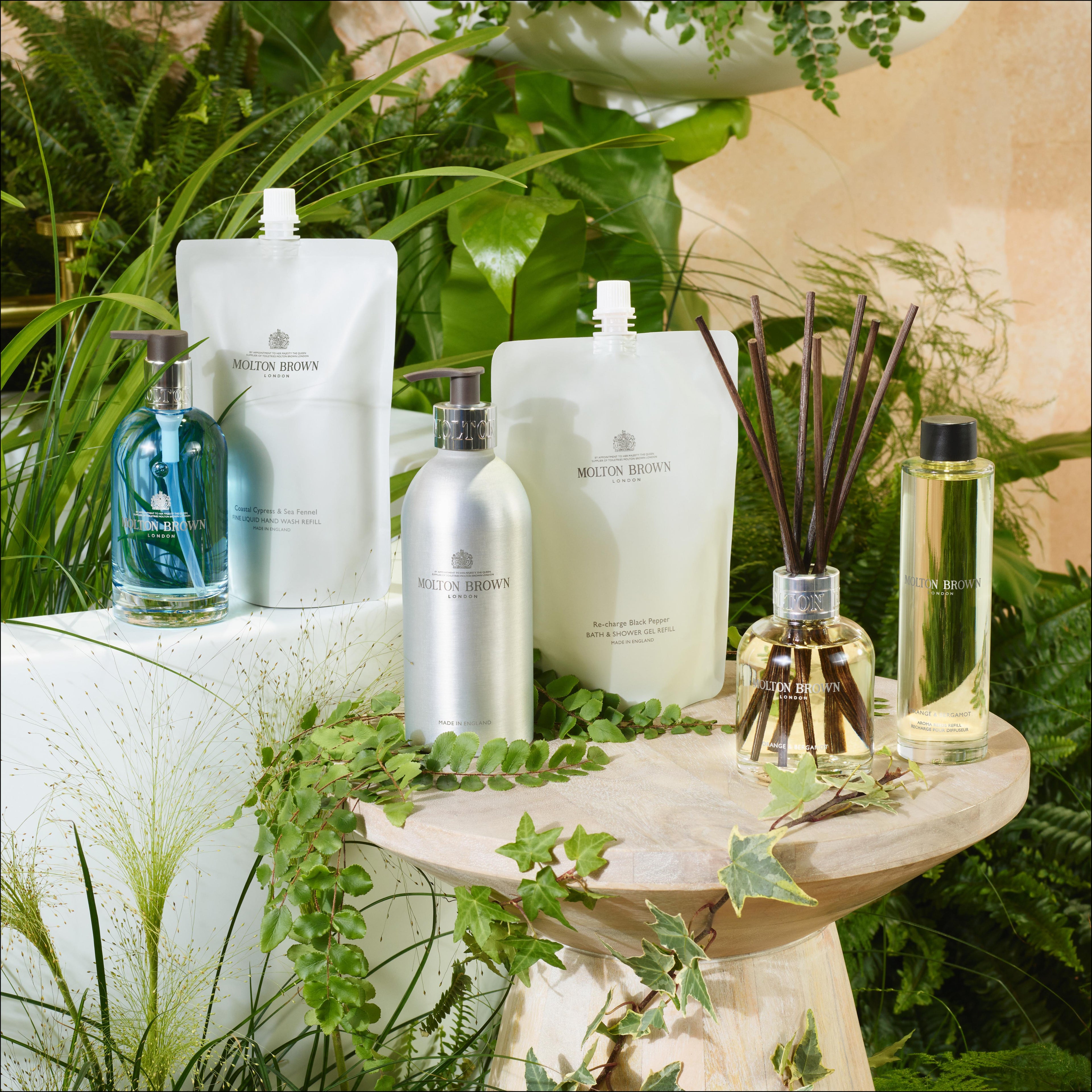 A display of MoltonBrown Cyprus Coastal Cypress & Sea Fennel Infinite Bottle 400ml is elegantly arranged on a wooden stool, featuring blue and metallic recycled aluminium bottles, refill pouches, and a reed diffuser. The lush greenery surrounding the setup creates a fresh and natural ambiance ideal for an eco-friendly bathing routine.