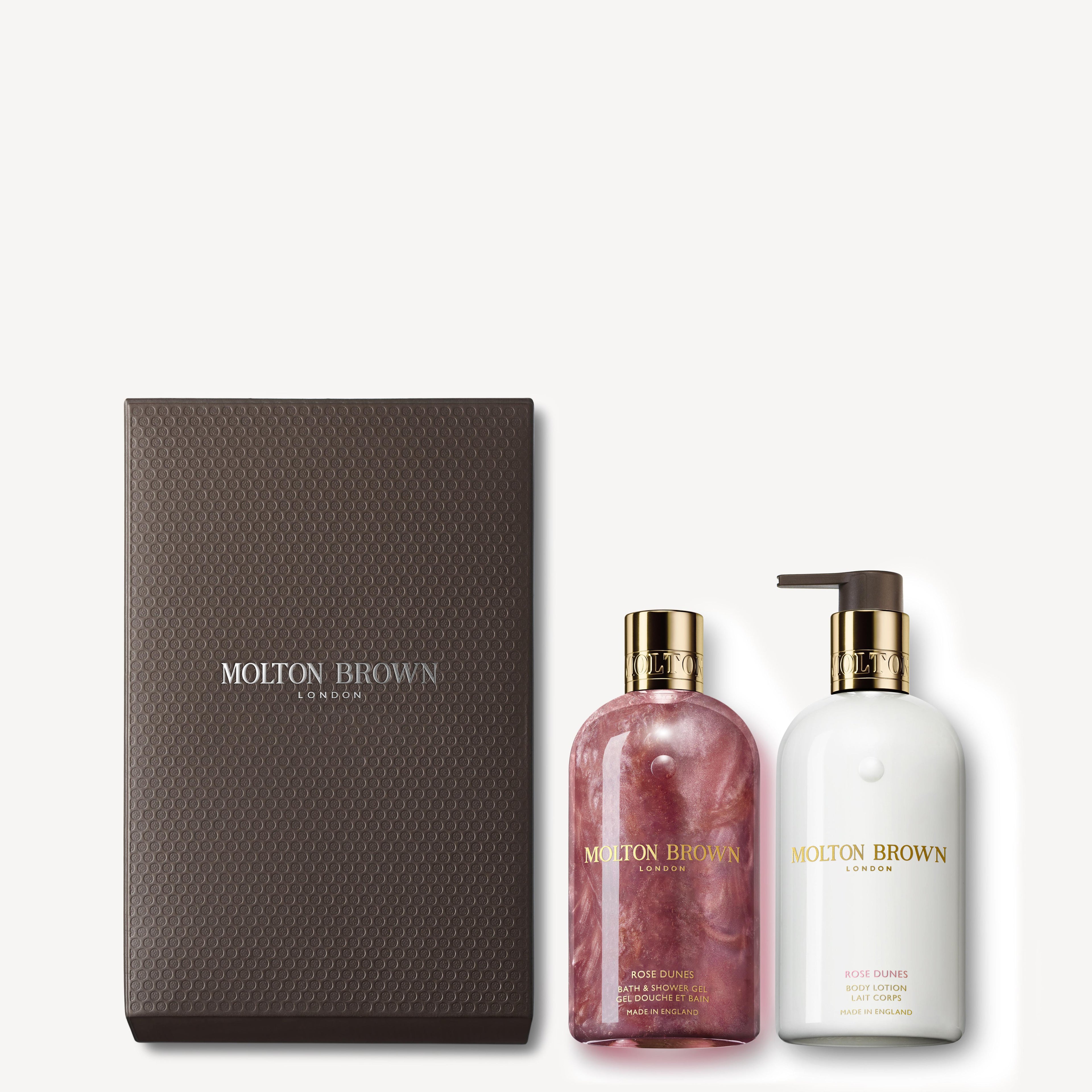 The Rose Dunes Body Care Gift Set by Molton Brown Cyprus includes a patchouli Sulawesi-infused pink body wash with a gold cap and a white lotion with a pump. Both are elegantly packaged in a dark brown box featuring the Molton Brown logo.