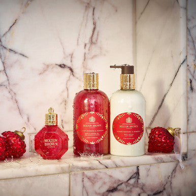 MoltonBrown Cyprus's Merry Berries & Mimosa Body Lotion sits elegantly on a marble surface, showcasing its luxurious red and white bottles with gold accents. Two glittery red heart decorations enhance the sophisticated spa-like atmosphere, making it ideal for nourishing winter skin with its floral-fruity aroma infused with the essence of red berries.