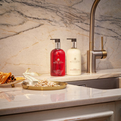 Two pump bottles, one red and one white, sit on a marble kitchen counter near a modern faucet. They are labeled MoltonBrown Cyprus, offering Festive Frankincense & Allspice Fine Liquid Hand Lotion. A decorative tray with dried botanicals sits to the left, adding charm to the elegant marble backsplash.