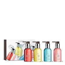 The Fresh & Floral Hand Care Collection from Molton Brown Cyprus features four luxury hand wash bottles in vibrant red, yellow, blue, and pink with pump dispensers. They are elegantly arranged before a white box with matching graphics and exude fruity-floral fragrances.