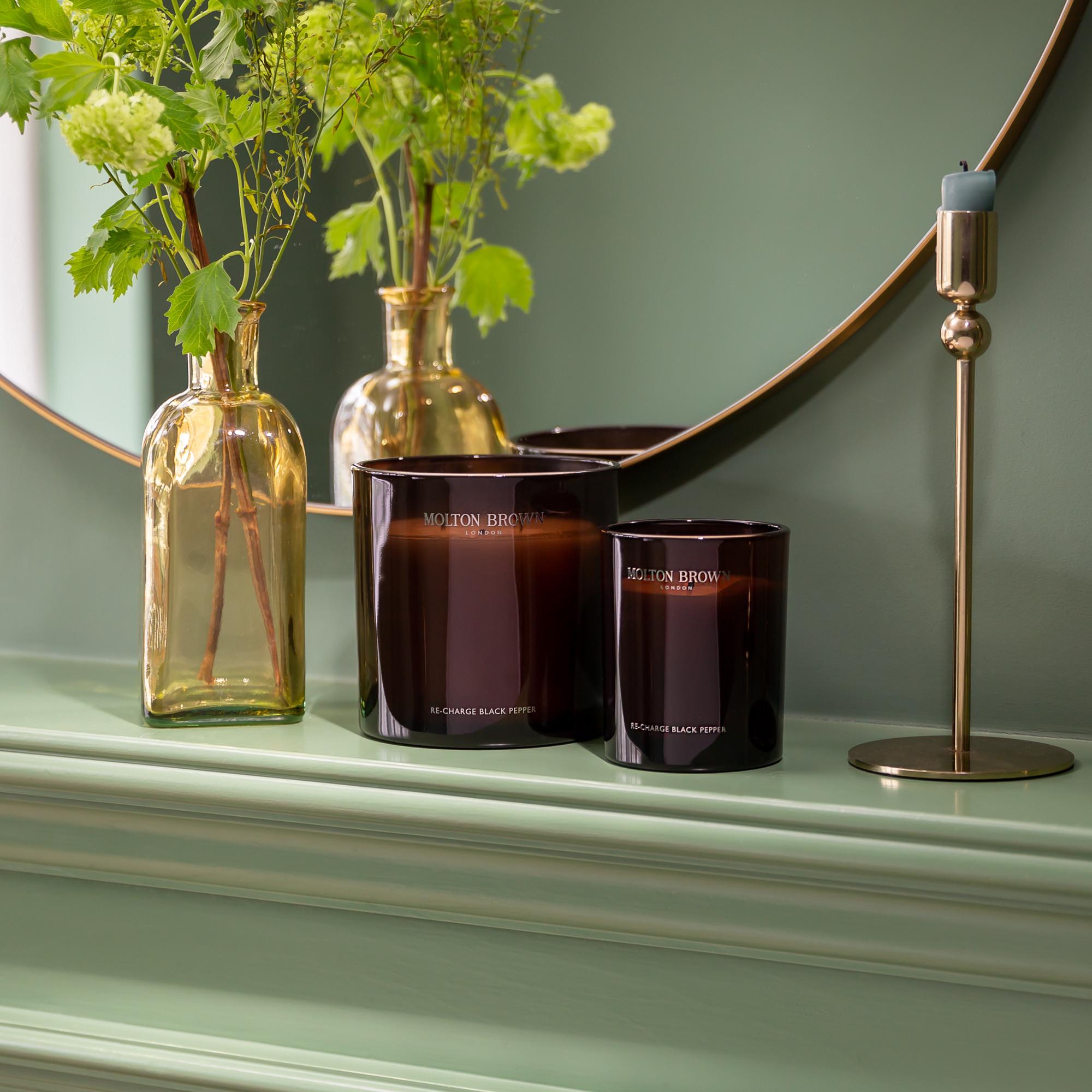 A serene green fireplace mantle displays two brown candles beside a golden vase with green foliage. A round mirror reflects the decor, while the Re-charge Black Pepper Signature Candle by MoltonBrown Cyprus, housed in a brass holder, adds elegance and rejuvenates the senses to this harmonious arrangement.