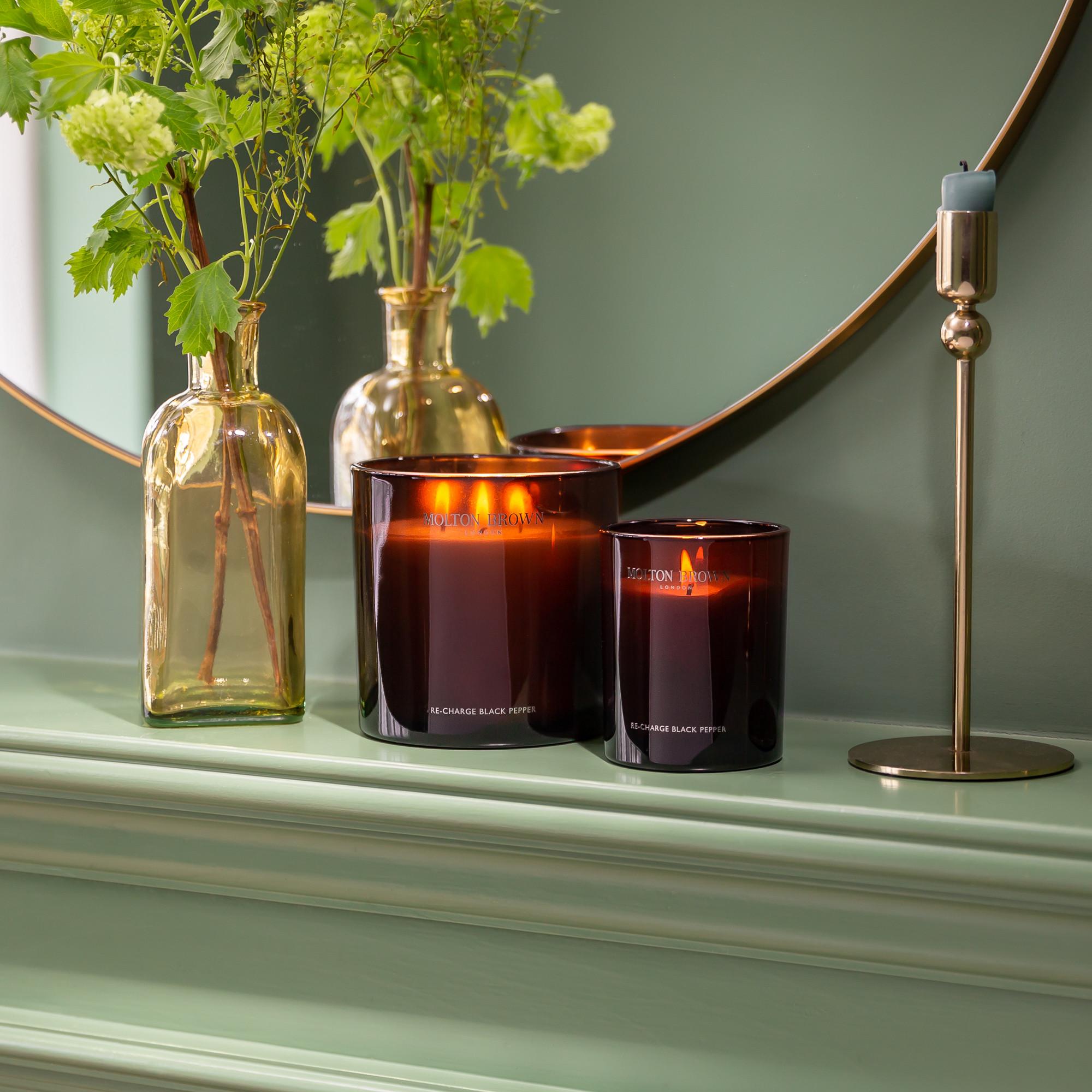 Two *Molton Brown* *Re-charge Black Pepper Signature Candles* are lit on a mint-green mantel. A round mirror reflects the gentle glow, filling the room with their rejuvenating Madagascan black pepper scent. A gold vase with green leaves and a tall candlestick holding an unlit candle contribute to the elegant and cozy atmosphere.