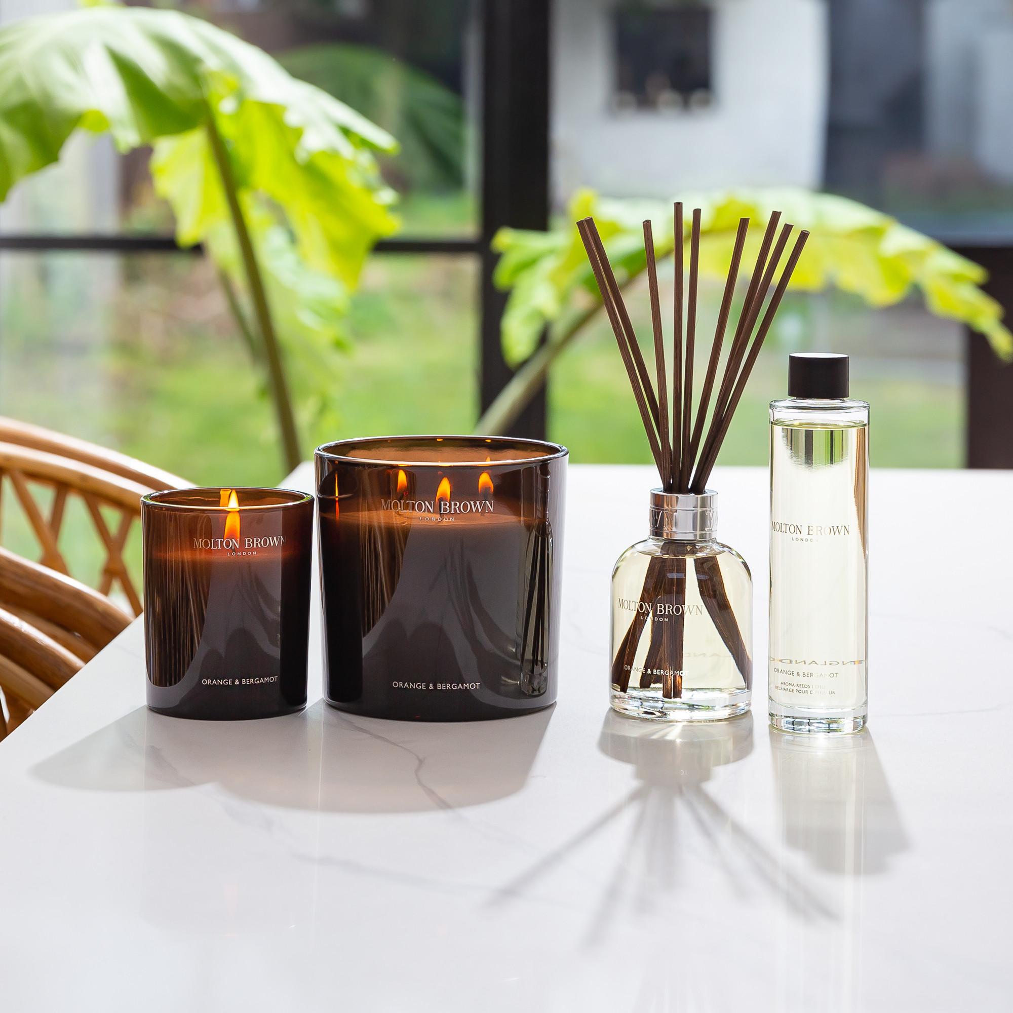 In a bright, airy room, the MoltonBrown Cyprus Orange & Bergamot Luxury Candle 600g and accompanying reed diffuser are elegantly displayed on a white table. The citrus white-floral fragrance enhances the calm ambiance, seamlessly blending with large green leaves, wicker furniture, and a window. This elegant space is thoughtfully crafted from recycled materials.