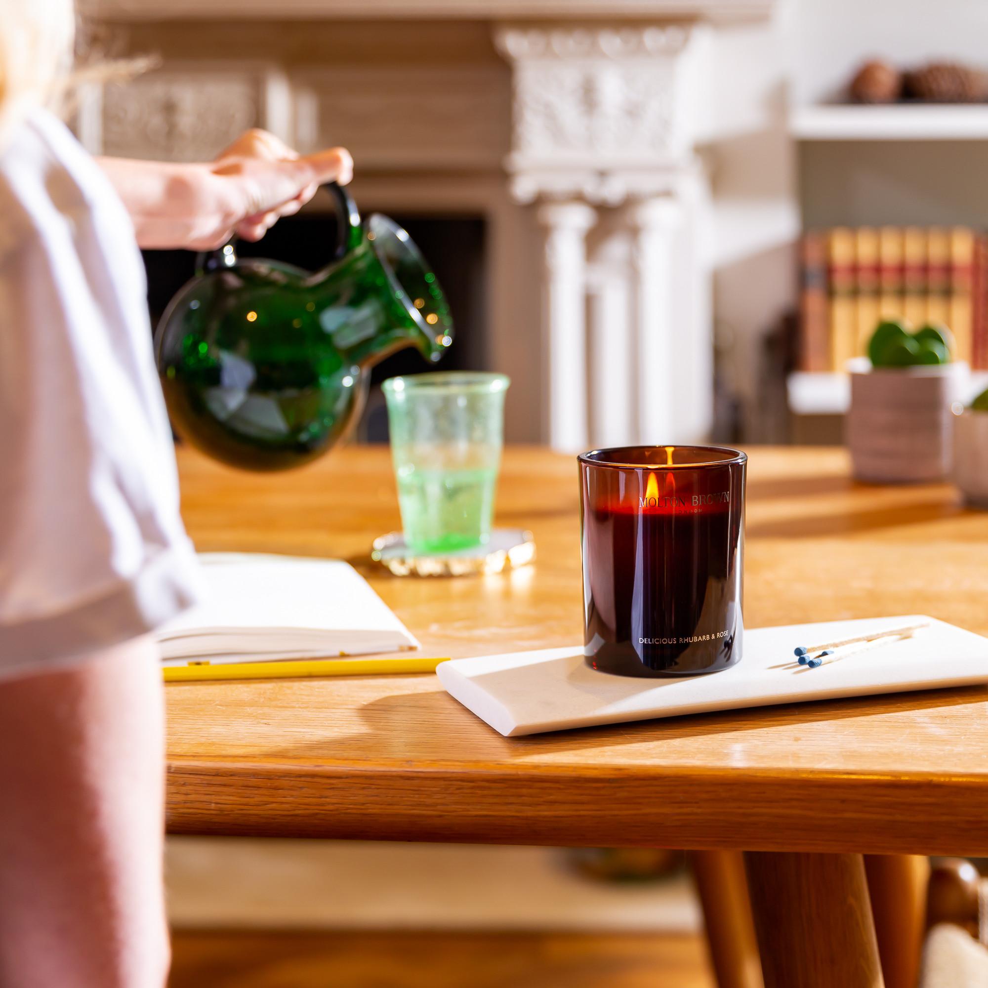 A person pours water from a green glass jug into a transparent cup on a wooden table, where the Delicious Rhubarb & Rose Signature Candle by MoltonBrown Cyprus gently flickers beside a notebook with a pen. A fireplace and bookshelves complete the cozy scene, infusing the air with its soothing fragrance.