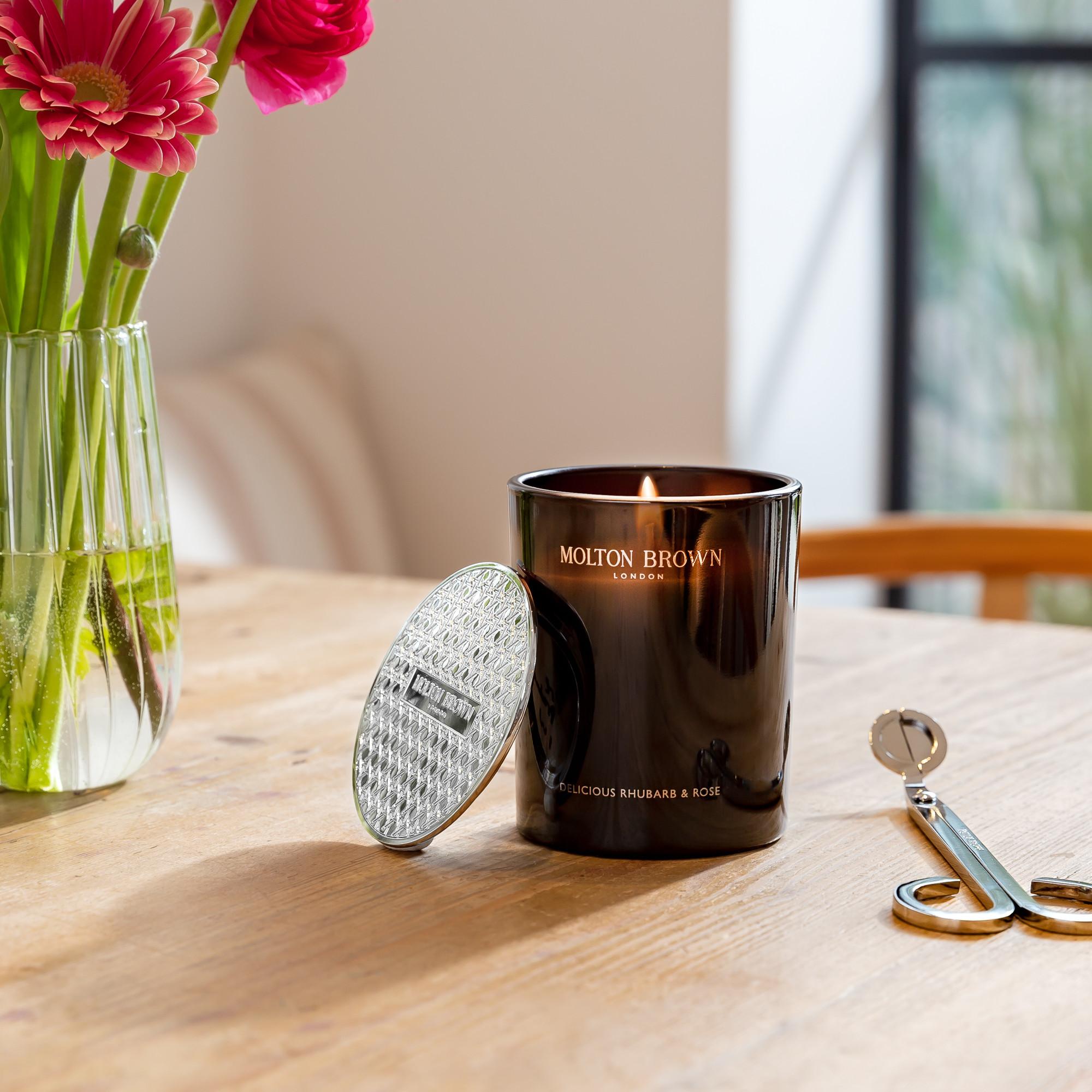 A lit Delicious Rhubarb & Rose Signature Candle 190g by MoltonBrown Cyprus rests on a wooden table with its lid beside it. Nearby are a pair of wick trimmers and a vase with vibrant pink flowers, filling the room with a floral-fruity fragrance. The ambiance is cozy and elegant.