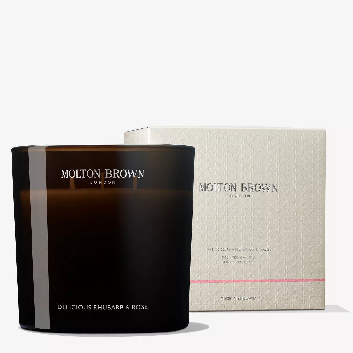 A luxury candle from MoltonBrown Cyprus, named Delicious Rhubarb & Rose Luxury Candle 600g, is showcased next to its sleek white packaging featuring understated patterns. This floral-fruity scent with a touch of grapefruit is housed in a glossy dark holder.