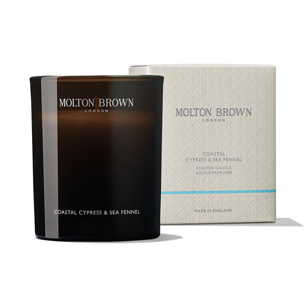 A MoltonBrown Cyprus Coastal Cypress & Sea Fennel Signature Candle 190g, housed in a dark glass container, sits next to its box. The candle's label indicates it is infused with marine notes. The light-colored box shares similar branding and design, embodying tranquil oceanside elegance.