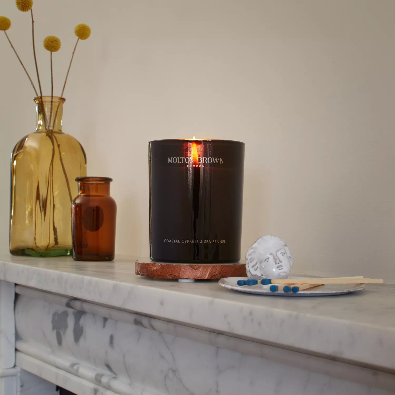 A lit Coastal Cypress & Sea Fennel Signature Candle by MoltonBrown Cyprus rests on a marble mantelpiece beside two brown glass bottles. A small decorative dish holds matchsticks and a white decorative object, while the fresh spices of the candle infuse the scene with a cozy and elegant ambiance.