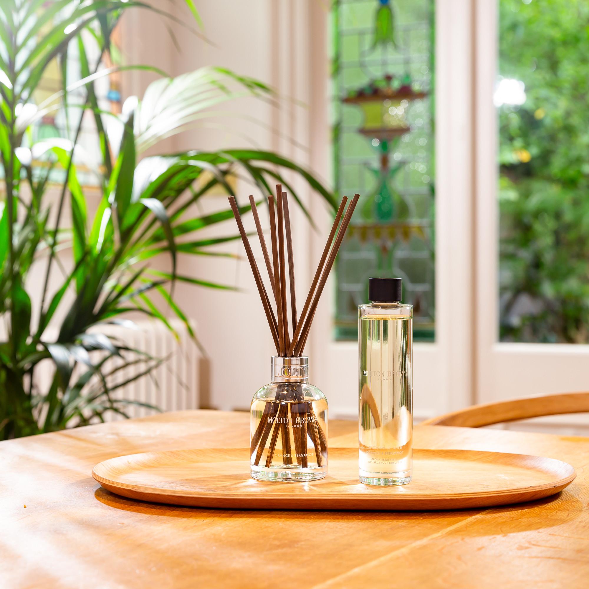 A wooden tray on a table holds a glass reed diffuser alongside a MoltonBrown Cyprus Orange & Bergamot Aroma Reeds Refill 150ml bottle, with green plants and a stained glass window in the background. The citrus home fragrance of Sevillian orange fills the air as bright natural light illuminates the serene scene.