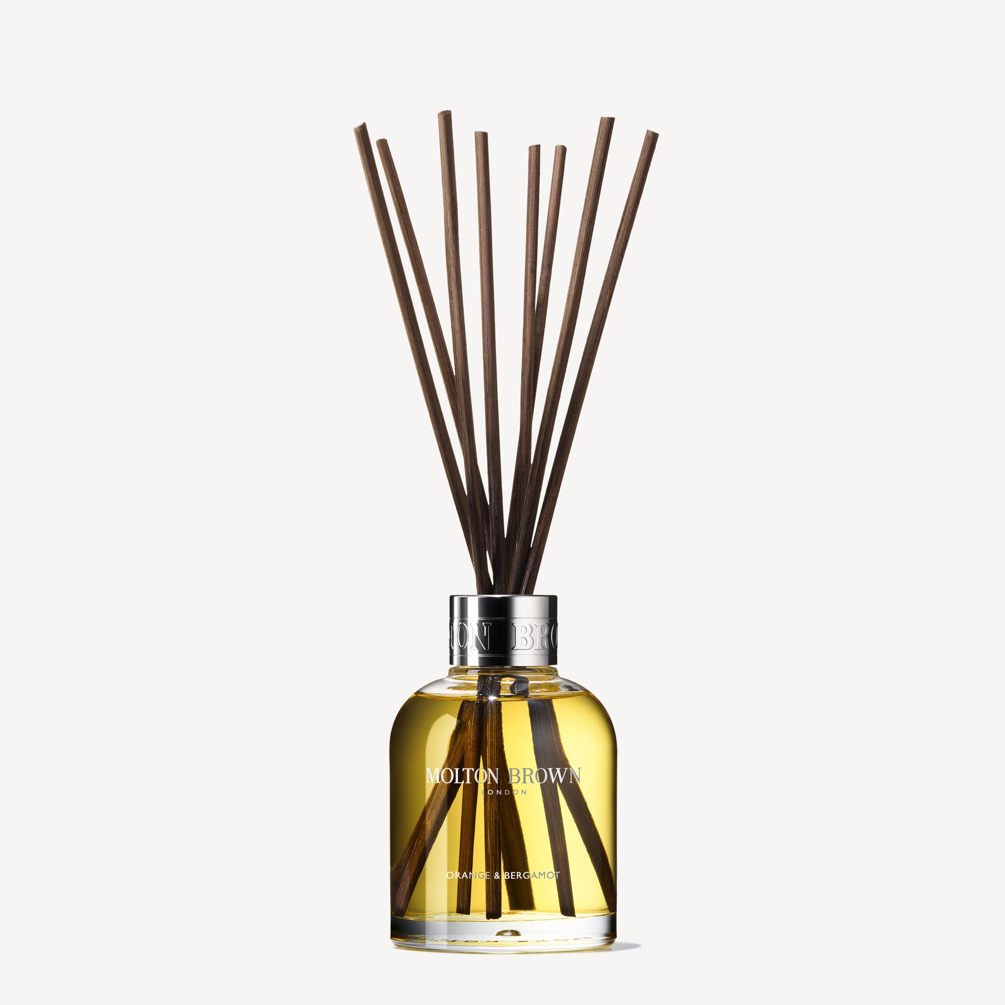 A glass bottle filled with a golden liquid, labeled MoltonBrown Cyprus, features dark reed sticks protruding from the top and serves as the Orange & Bergamot Aroma Reeds 150ml diffuser. The light off-white backdrop accentuates its elegance.