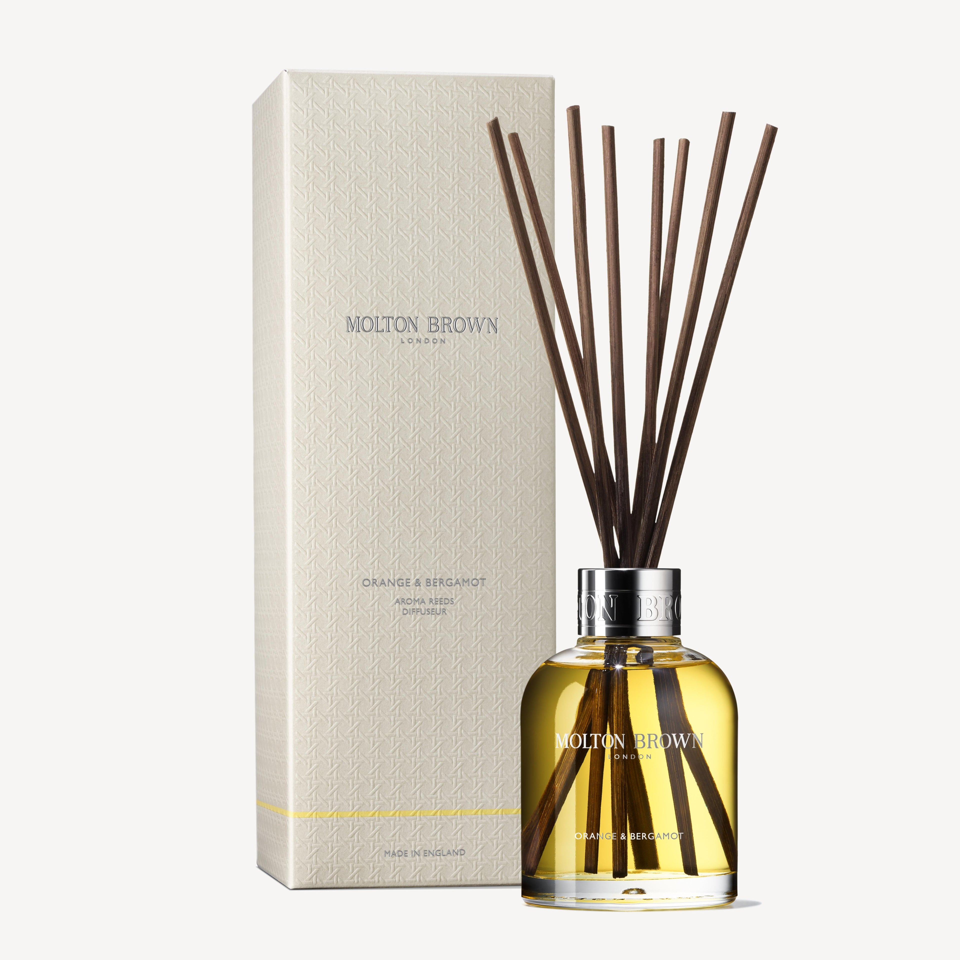 The scene is enhanced by a MoltonBrown Cyprus Orange & Bergamot Aroma Reeds 150ml diffuser, featuring a clear bottle filled with a yellow liquid and dark reeds. Next to it, a textured cream box unveils the sophisticated aroma of Sevillian orange and neroli blooms, exuding elegance and aromatic charm.