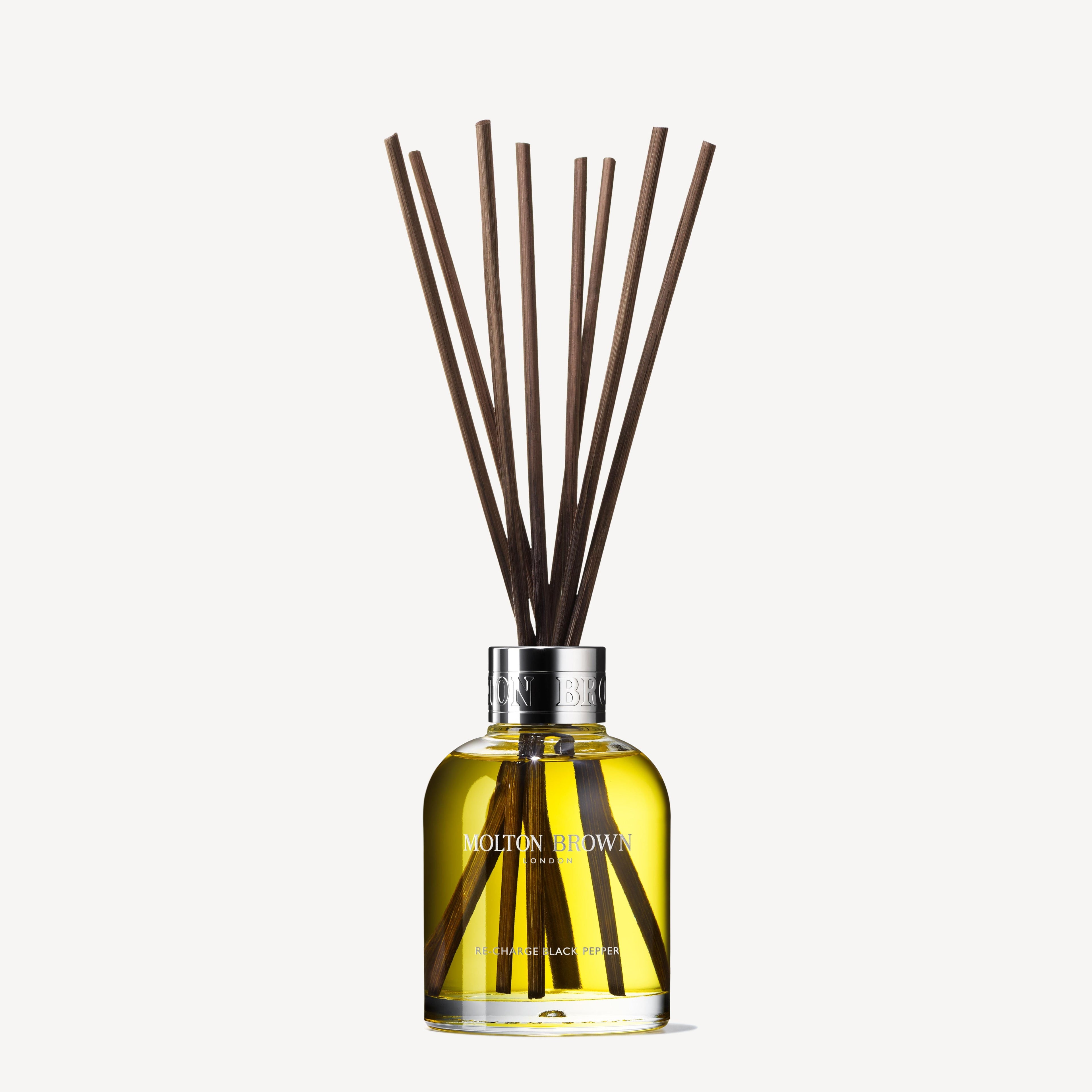 A glass bottle of MoltonBrown Cyprus Re-charge Black Pepper Aroma Reeds with dark reed sticks elegantly emerging from the top. The vibrant yellow oil exudes a woody-citrus aroma, and the label displays MoltonBrown Cyprus with details of Madagascan black pepper below. The background is plain white.