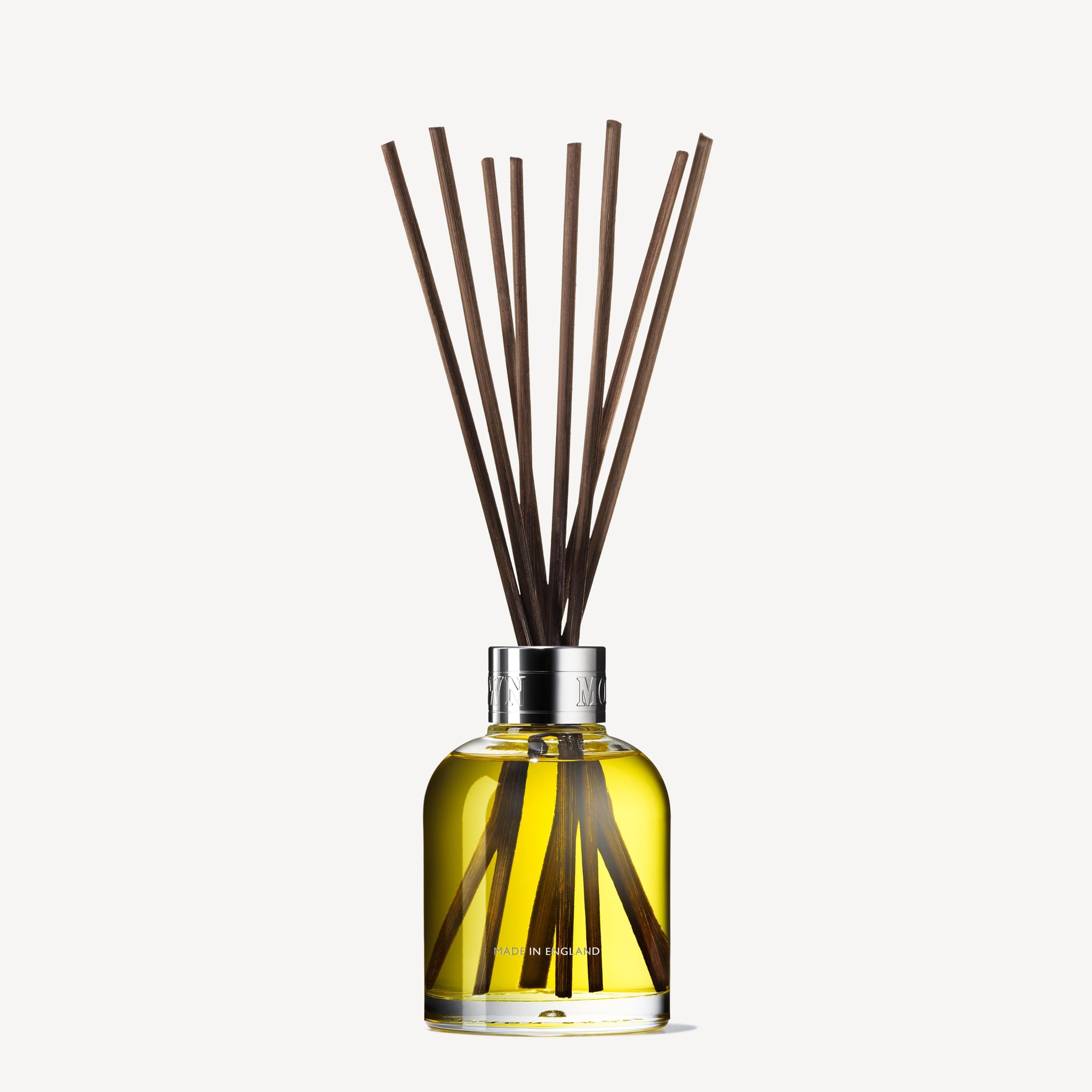 The Re-charge Black Pepper Aroma Reeds by MoltonBrown Cyprus feature a glass bottle filled with yellow liquid, topped with a shiny silver cap, and contains several long reed sticks protruding upwards. These reeds diffuse a refreshing woody-citrus fragrance, infused with hints of vetiver, into the surrounding space.