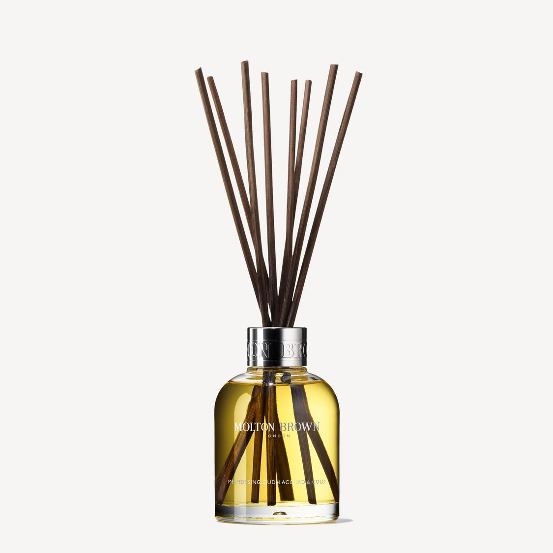 A clear glass bottle filled with yellow Mesmerising Oudh Accord & Gold fragrance by MoltonBrown Cyprus, topped with a silver cap and several long, dark brown aroma reeds fanned at the top. 150ml of scented decadence awaits inside.
