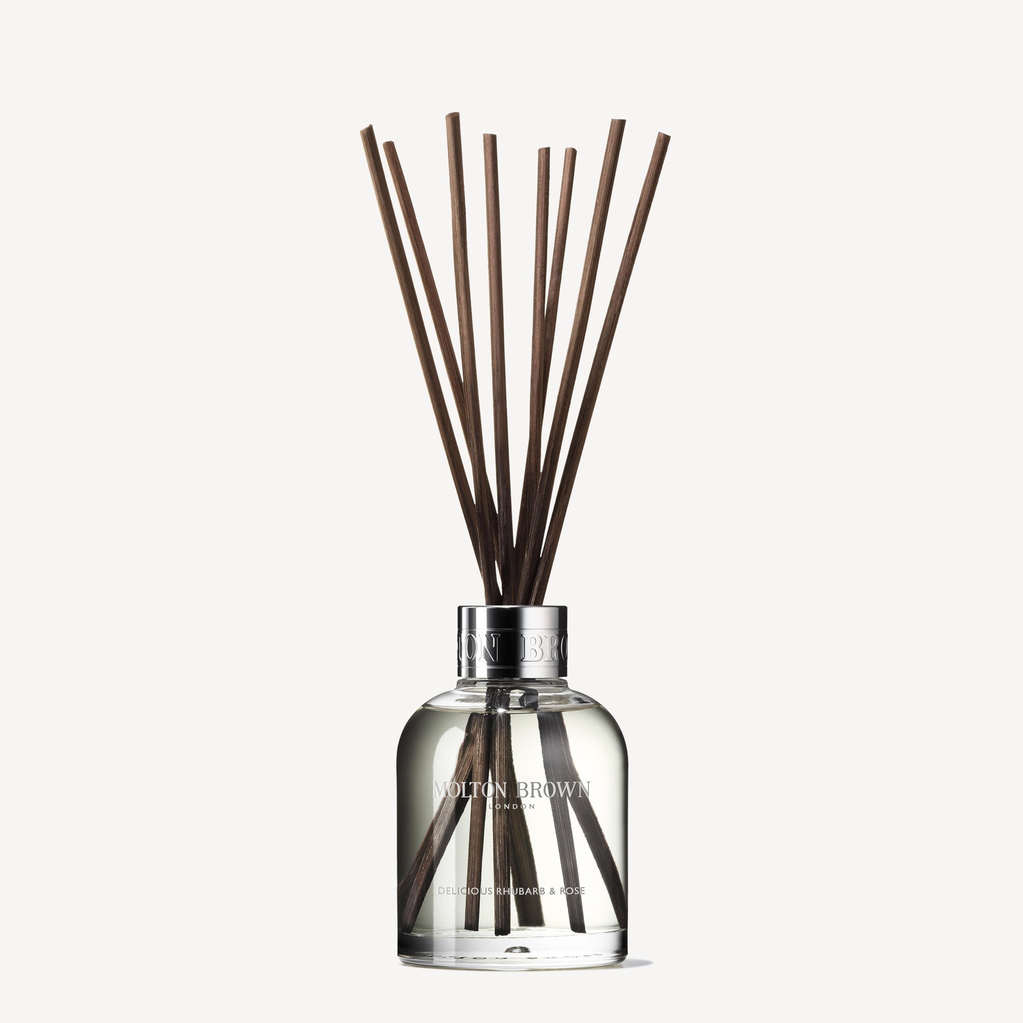 The Delicious Rhubarb & Rose Aroma Reeds 150ml from MoltonBrown Cyprus comes in a clear glass bottle, showcasing several long, dark brown reeds that extend upward. The visible liquid emits enticing exotic rhubarb and rose scents. A silver cap adorned with the brand name completes this sophisticated accessory, promising a luxurious aroma.