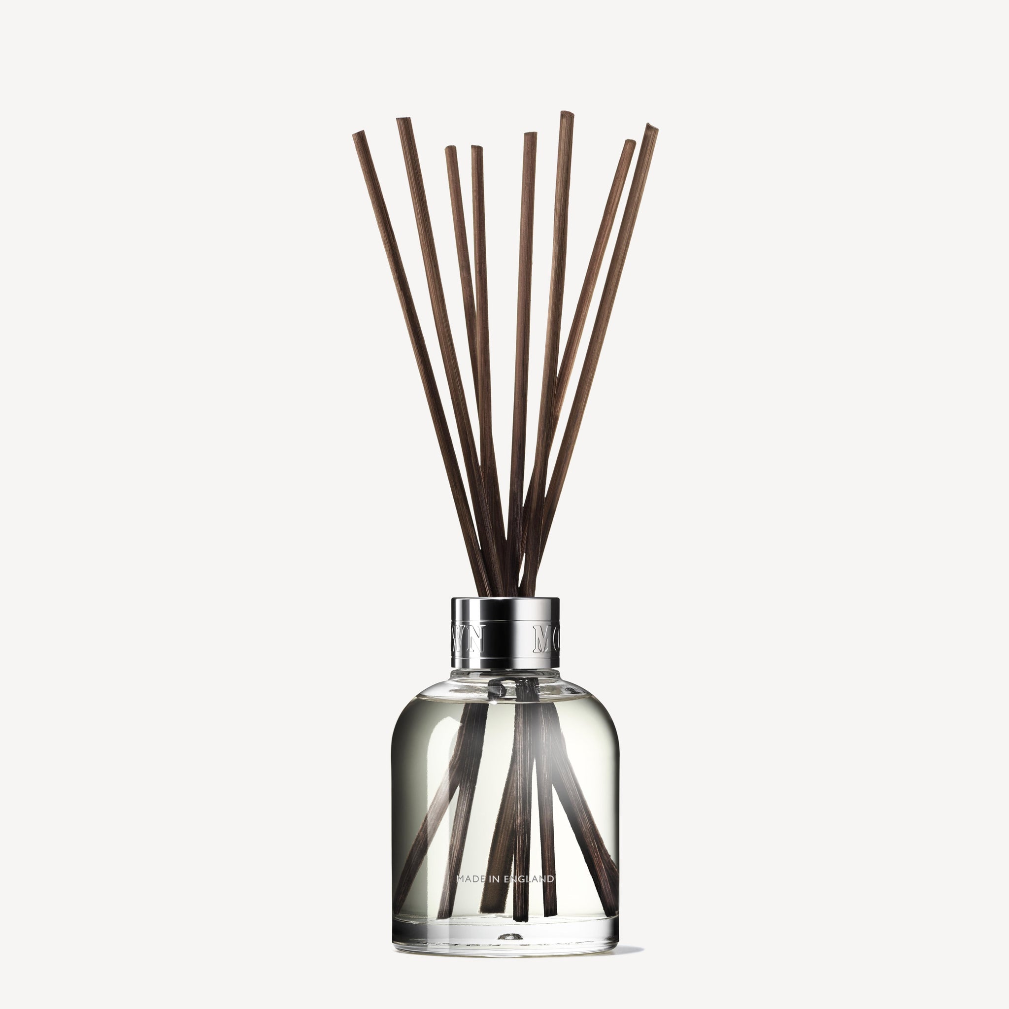 The MoltonBrown Cyprus Delicious Rhubarb & Rose Aroma Reeds 150ml is an elegant home fragrance solution, featuring a clear glass diffuser bottle filled with liquid that emits subtle rhubarb and rose scents. Topped with a silver cap, wooden reeds are arranged to fan out from above against a simple, light background.