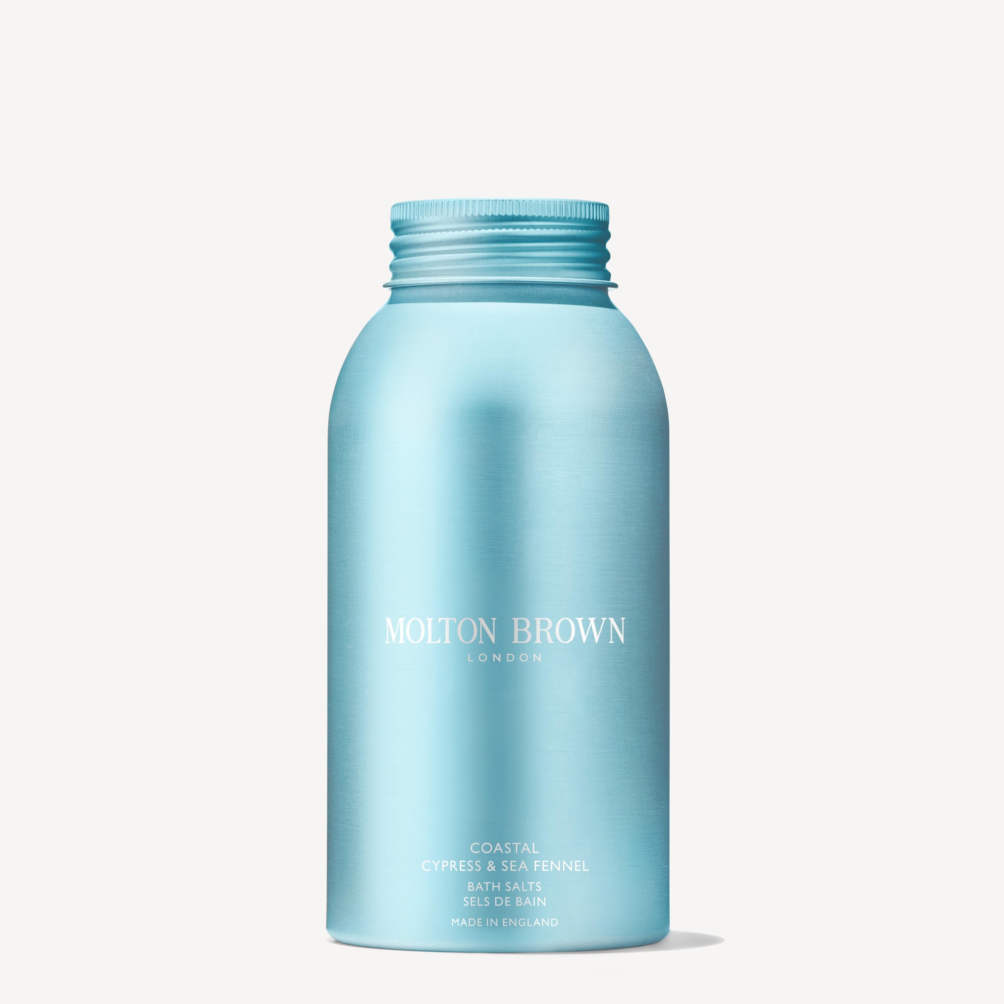 A hand holds a blue bottle of MoltonBrown Cyprus Coastal Cypress & Sea Fennel Bath Salts 300g above a partially filled bathtub with running water. The calming aroma of Coastal Cypress harmonizes beautifully with the bathroom's sleek chrome faucet fixtures, creating an inviting escape.