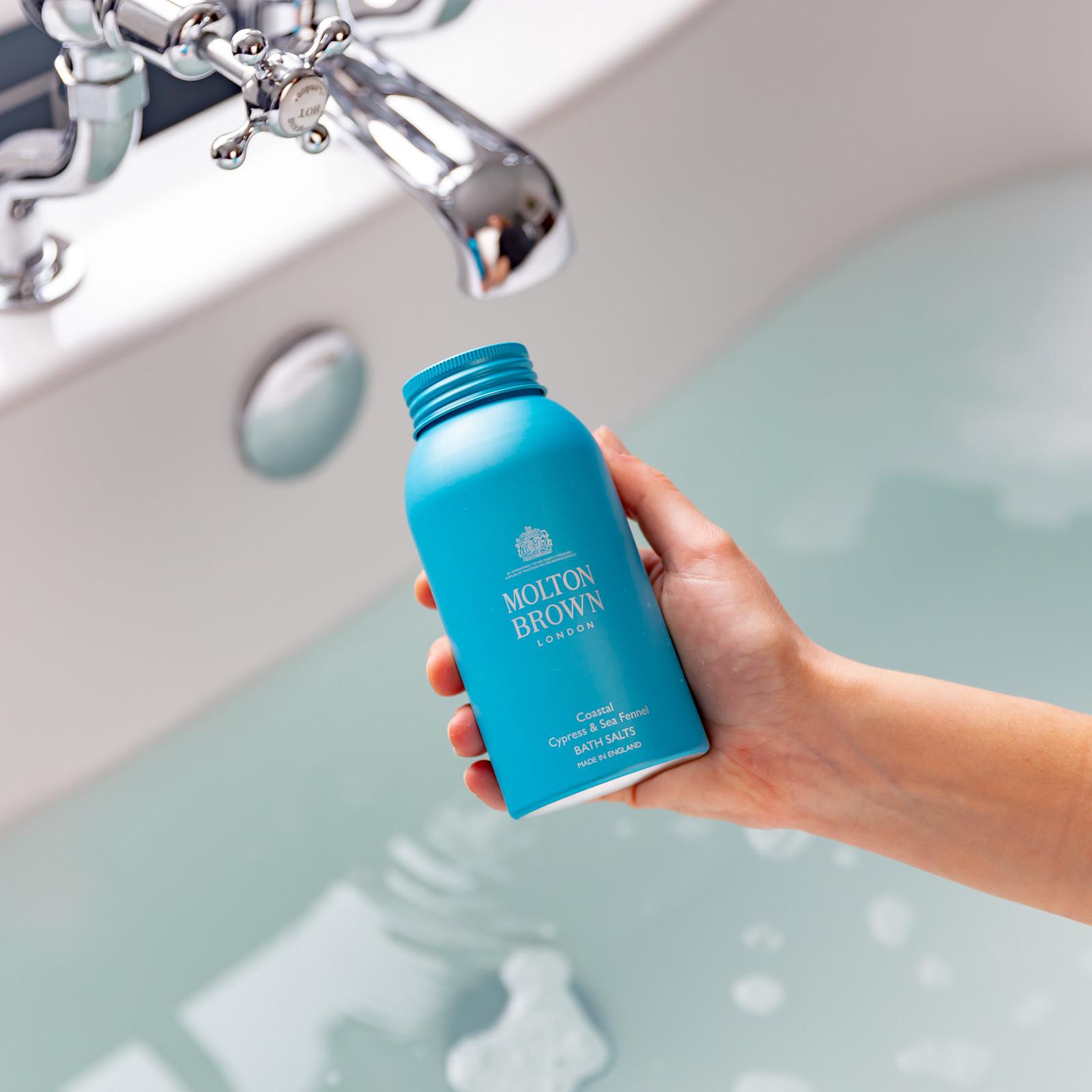 A hand holds a blue bottle of MoltonBrown Cyprus Coastal Cypress & Sea Fennel Bath Salts 300g above a partially filled bathtub with running water. The calming aroma of Coastal Cypress harmonizes beautifully with the bathroom's sleek chrome faucet fixtures, creating an inviting escape.