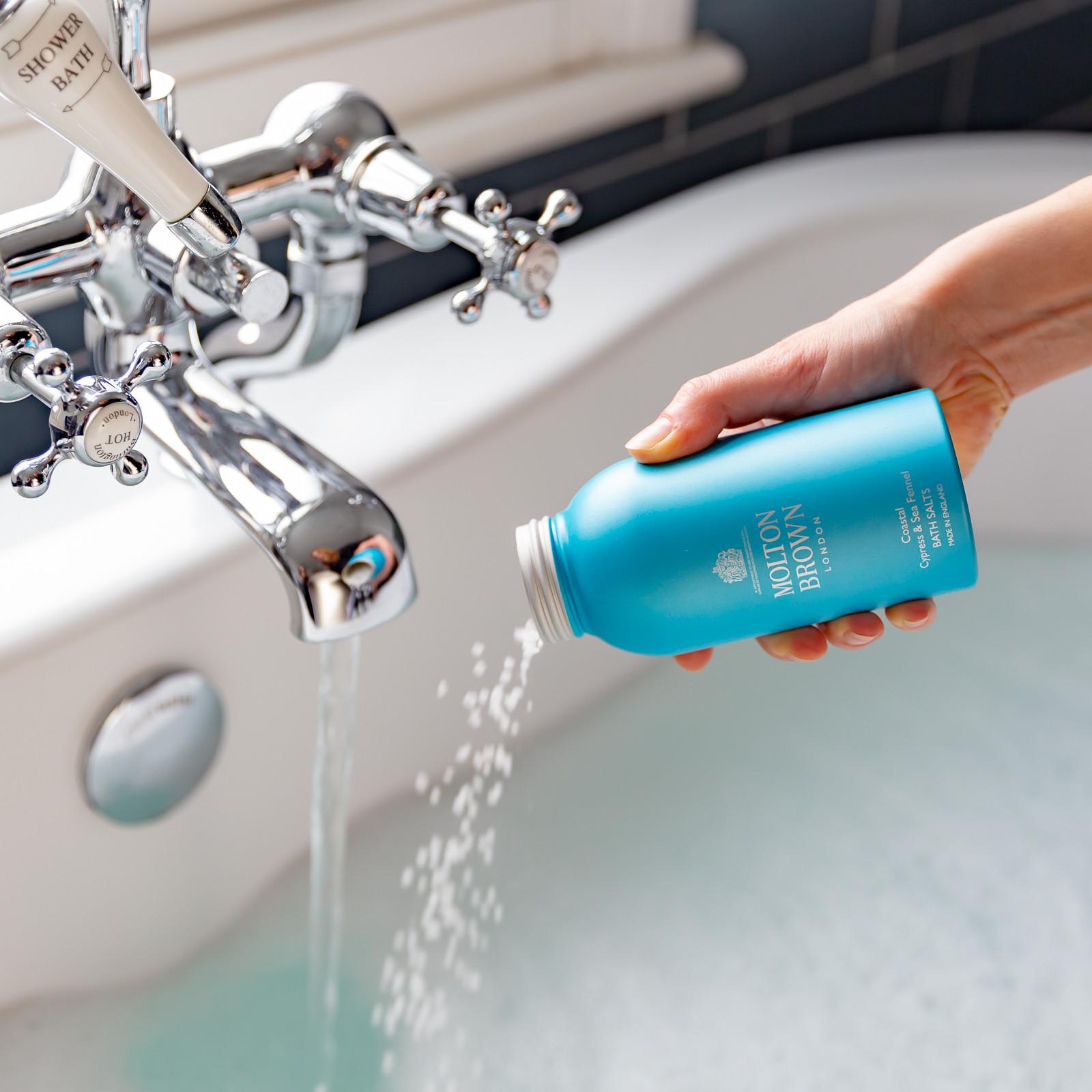 A hand pours bath salts from a 300g blue bottle of MoltonBrown Cyprus Coastal Cypress & Sea Fennel into a flowing bath faucet, forming bubbles in the bathtub below. The faucet is equipped with multiple handles, set against the backdrop of a serene bathroom.