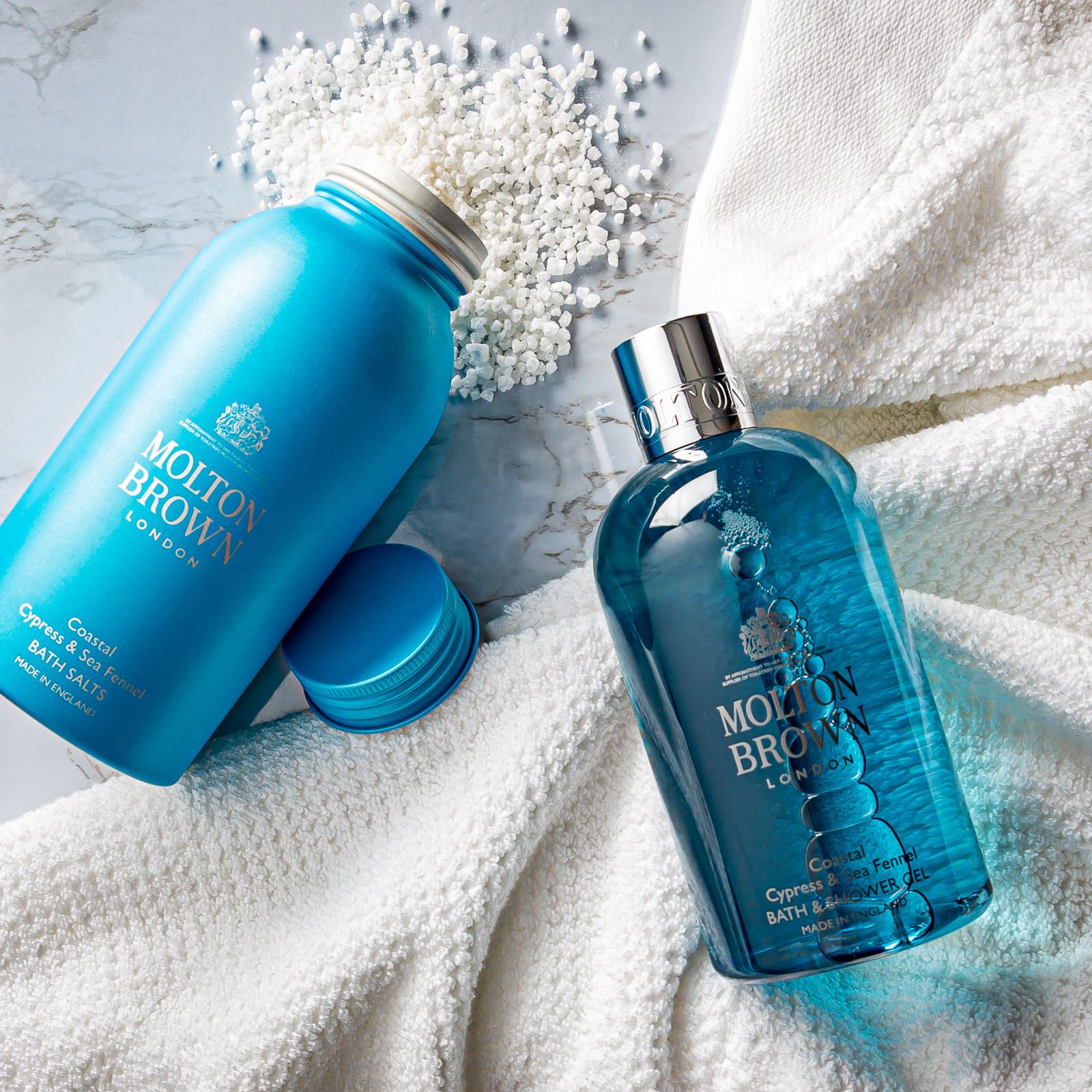 Two blue bottles of MoltonBrown Cyprus bath products are elegantly displayed on a light marble surface with a white textured towel. The Coastal Cypress & Sea Fennel Bath Salts, weighing 300g, are open with salts artfully scattered around one bottle, while the other remains closed, creating a luxurious and serene bath setting.