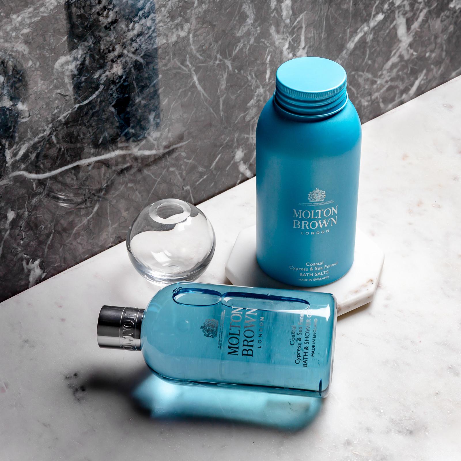 The MoltonBrown Cyprus bath product set includes a blue bath and shower gel bottle infused with Coastal Cypress, alongside a container of Coastal Cypress & Sea Fennel Bath Salts 300g on a marble surface, accompanied by a small decorative glass sphere.