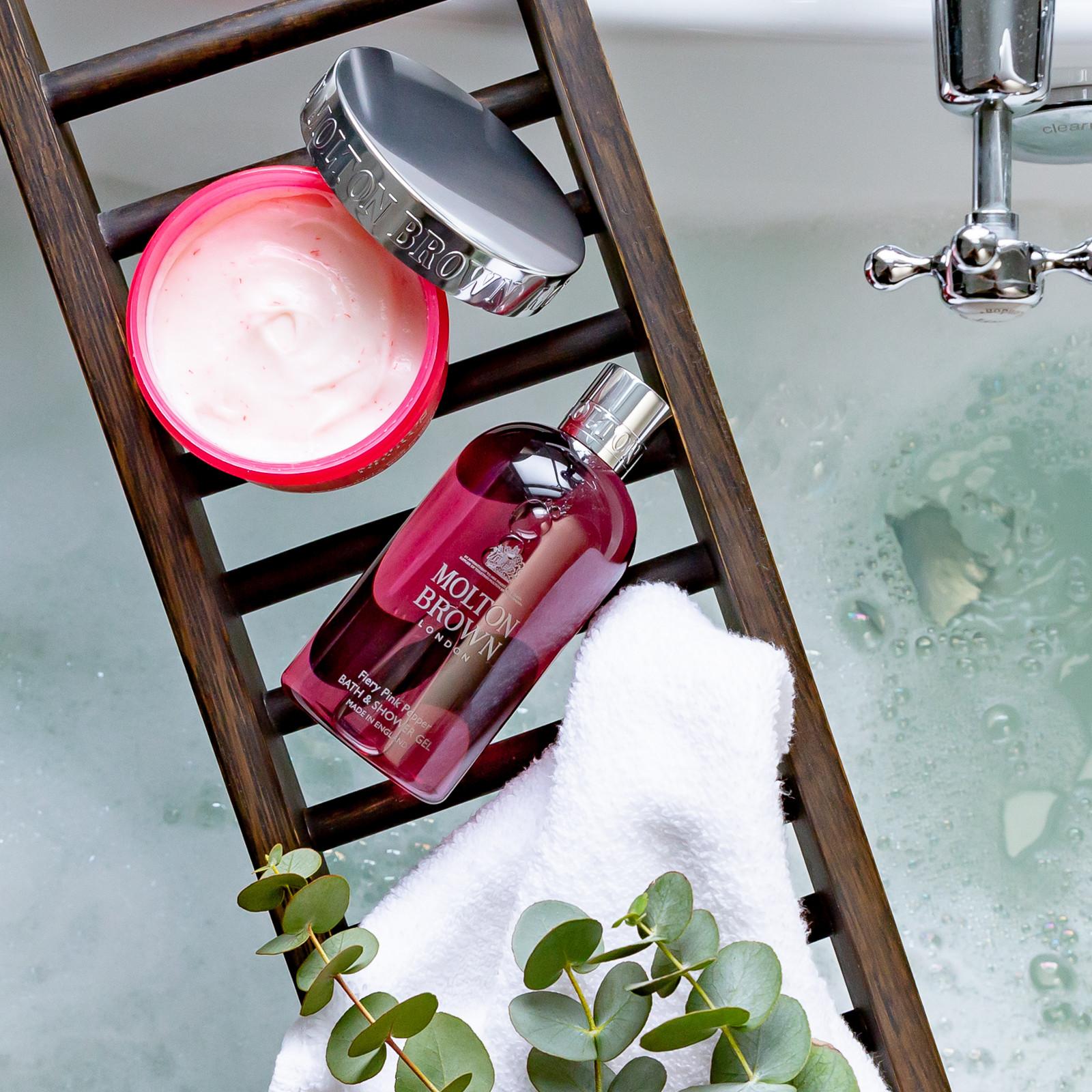 The bathtub caddy holds a pink jar of cream with a silver lid, a red bottle of MoltonBrown Cyprus Fiery Pink Pepper Pampering Body Polisher 275g, and an exfoliating bamboo body scrub. A fluffy white towel and fresh eucalyptus leaves add charm to the scene. In the background, a tub brims with water and bubbles.