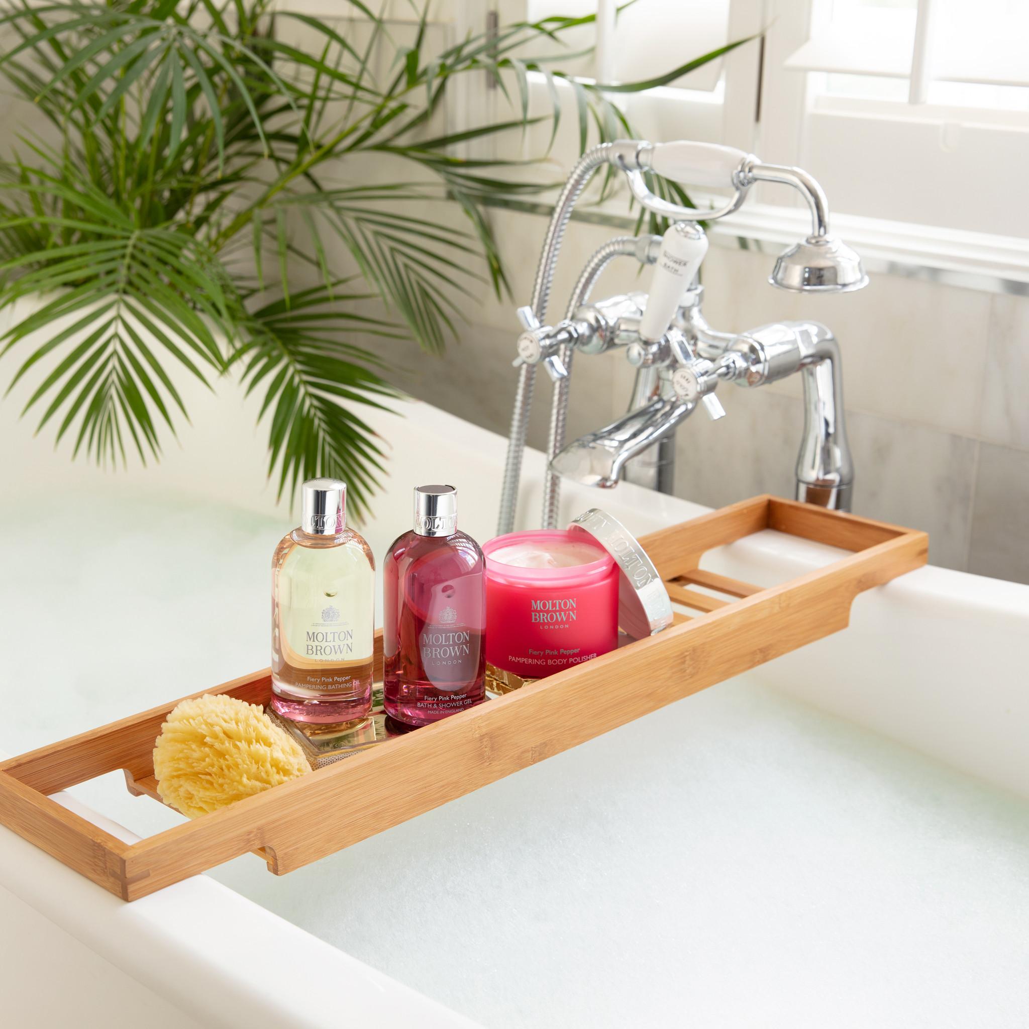 A bathtub with a wooden tray holding Molton Brown Cyprus Fiery Pink Pepper Pampering Body Polisher 275g, an exfoliating bamboo body scrub, a pink candle, and a bath sponge. The tub brims with water as a palm plant adds greenery near the window in the background.