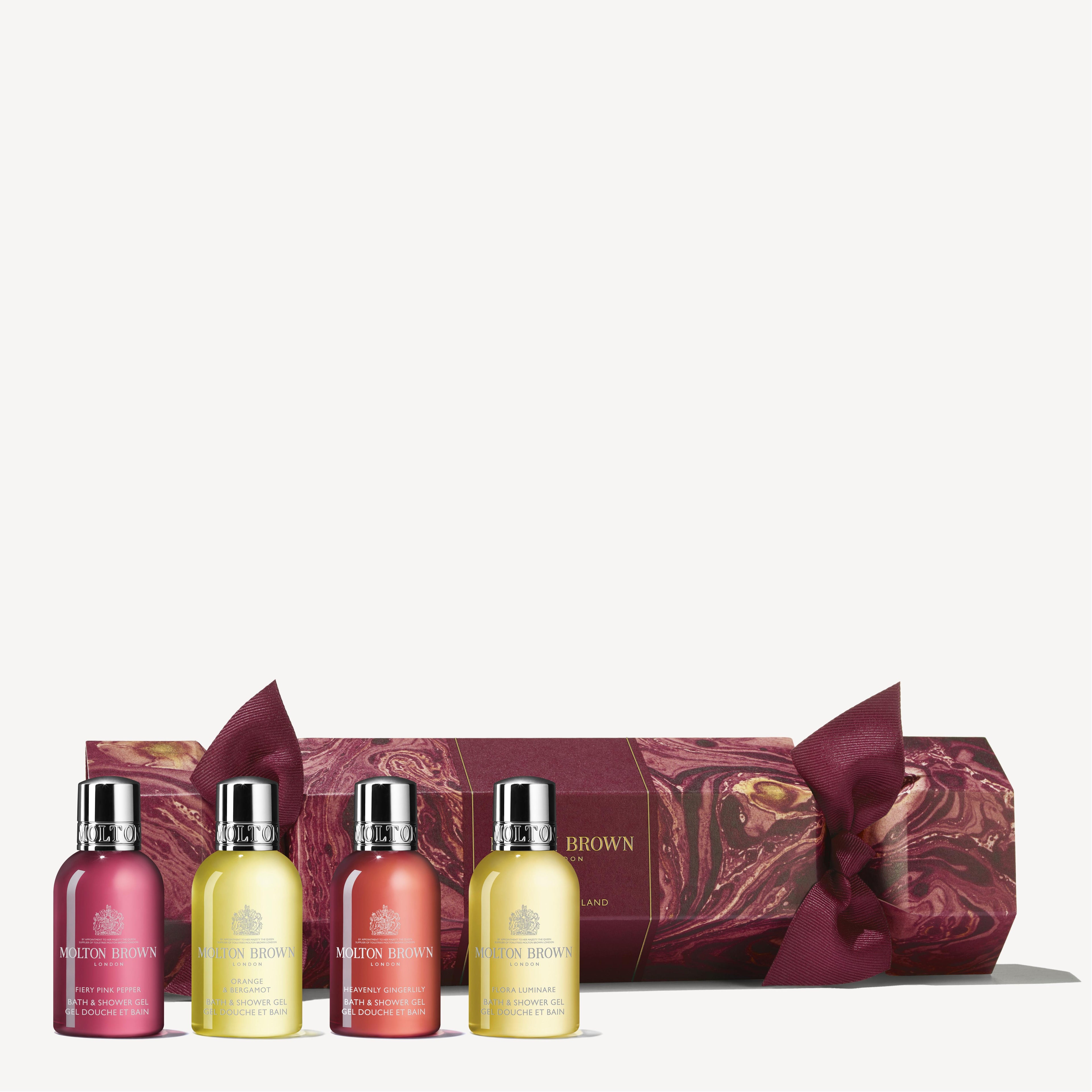 A set of four small bottles of MoltonBrown Cyprus shower gel in different colors is displayed in front of a maroon and gold patterned gift box, elegantly shaped like the Floral & Spicy Christmas Cracker, making it perfect for any Christmas table setting.