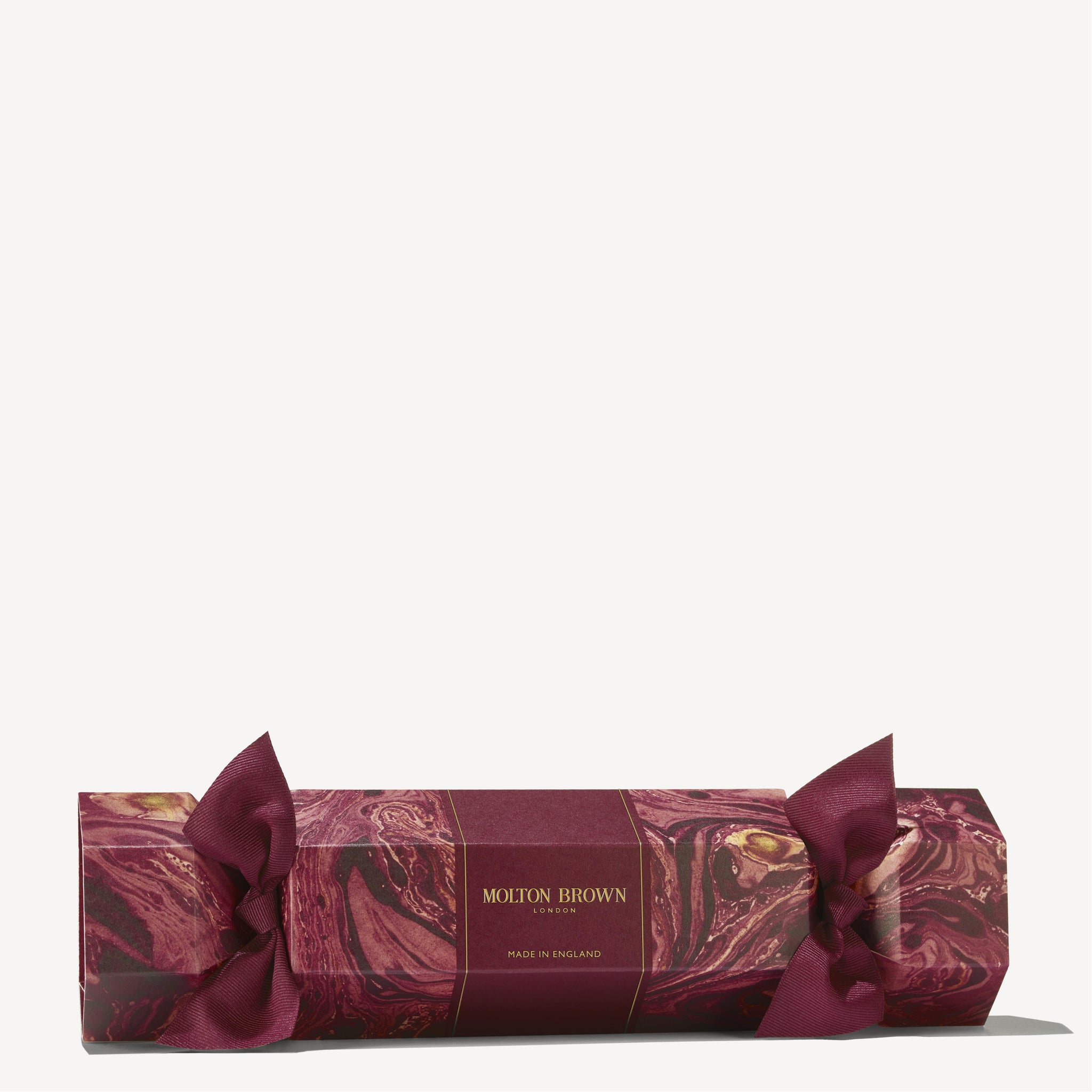 A Floral & Spicy Christmas Cracker from MoltonBrown Cyprus, featuring a maroon and gold gift box with a marble pattern and tied with maroon ribbons at both ends, resembles a festive cracker, making it ideal for embellishing your Christmas table against a light background.