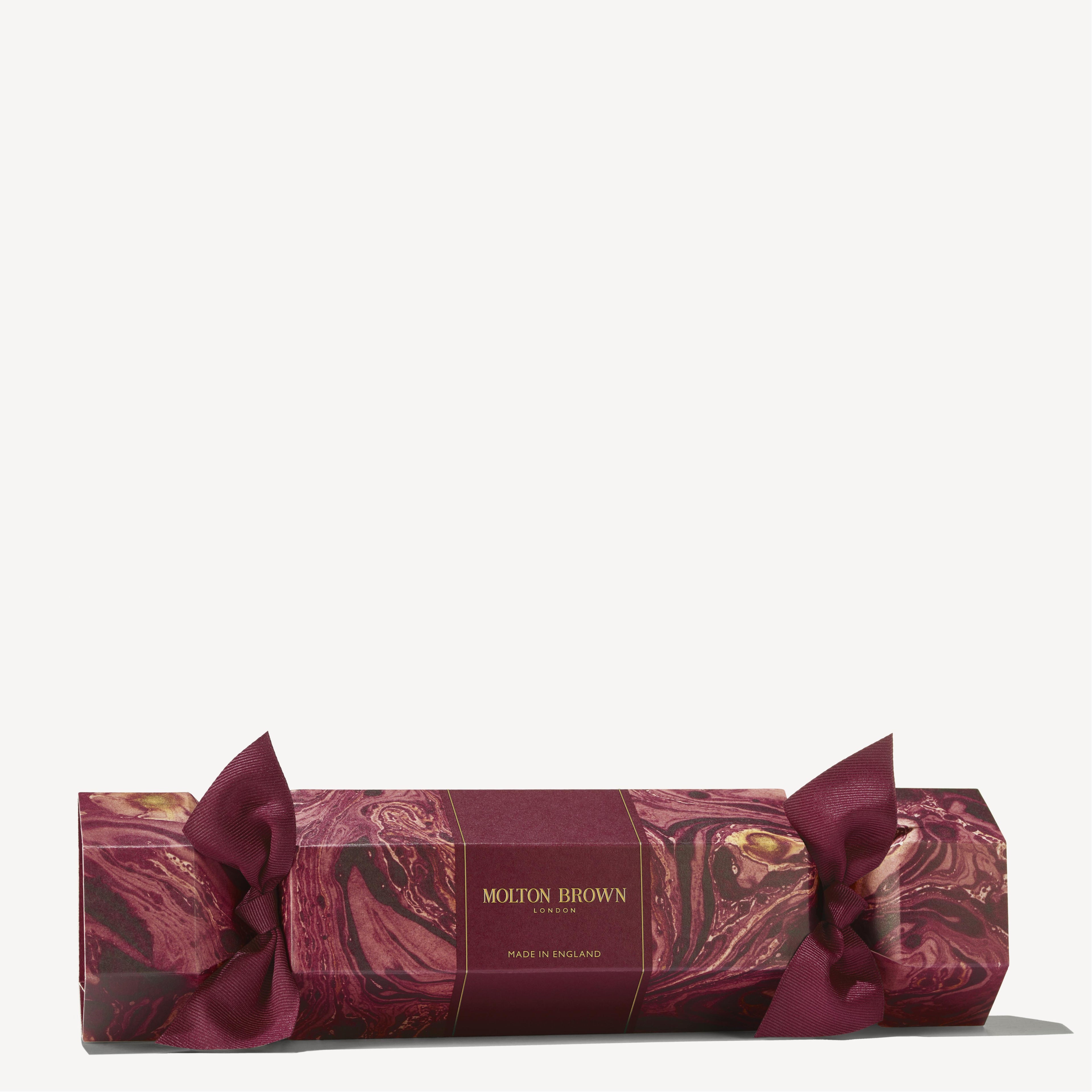 A Floral & Spicy Christmas Cracker from MoltonBrown Cyprus, featuring a maroon and gold gift box with a marble pattern and tied with maroon ribbons at both ends, resembles a festive cracker, making it ideal for embellishing your Christmas table against a light background.