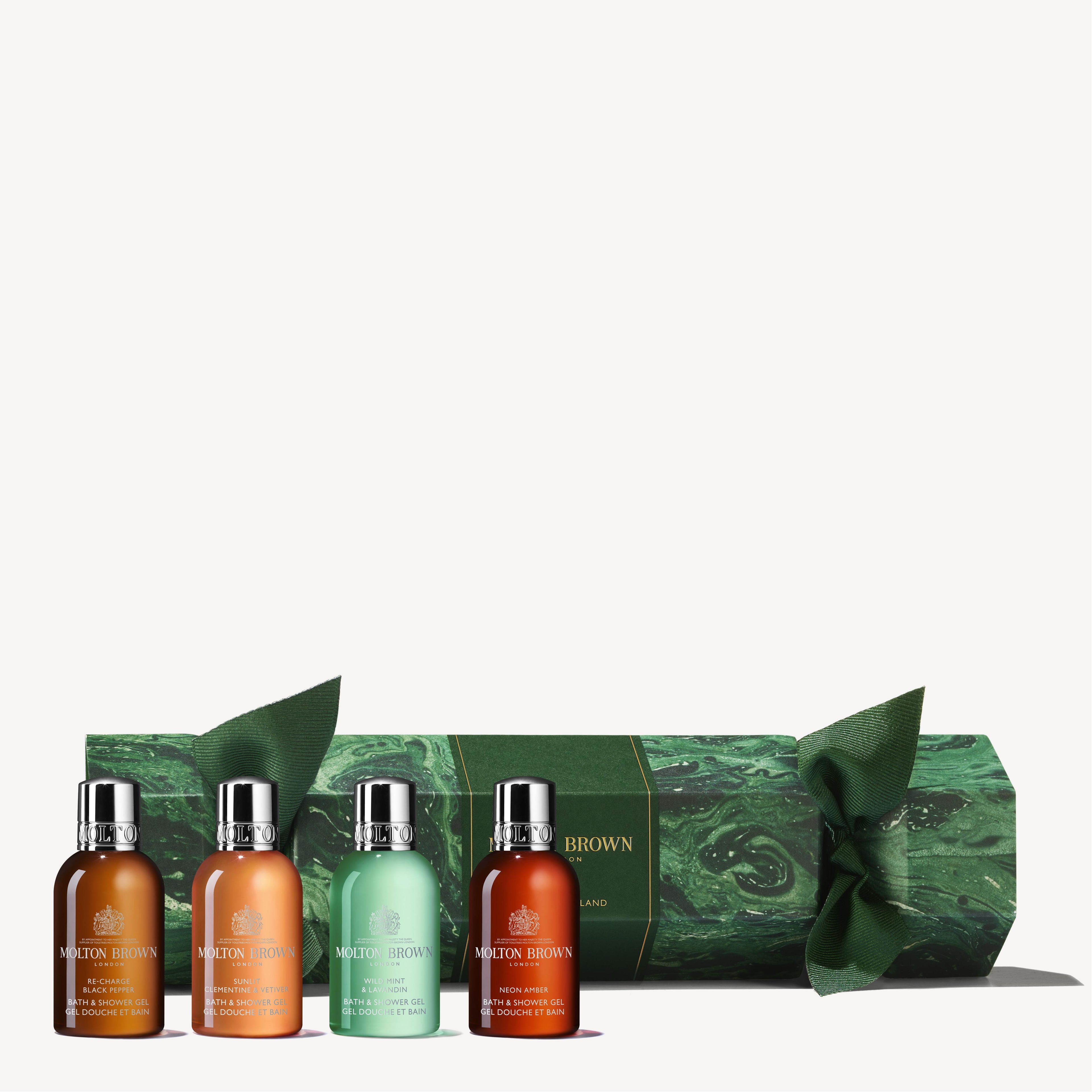 Arranged in front of the lushly designed green marble-patterned Woody & Aromatic Christmas Cracker with tied ends, four small bottles of MoltonBrown Cyprus's body washes sit lined up, each displaying vibrant colors.