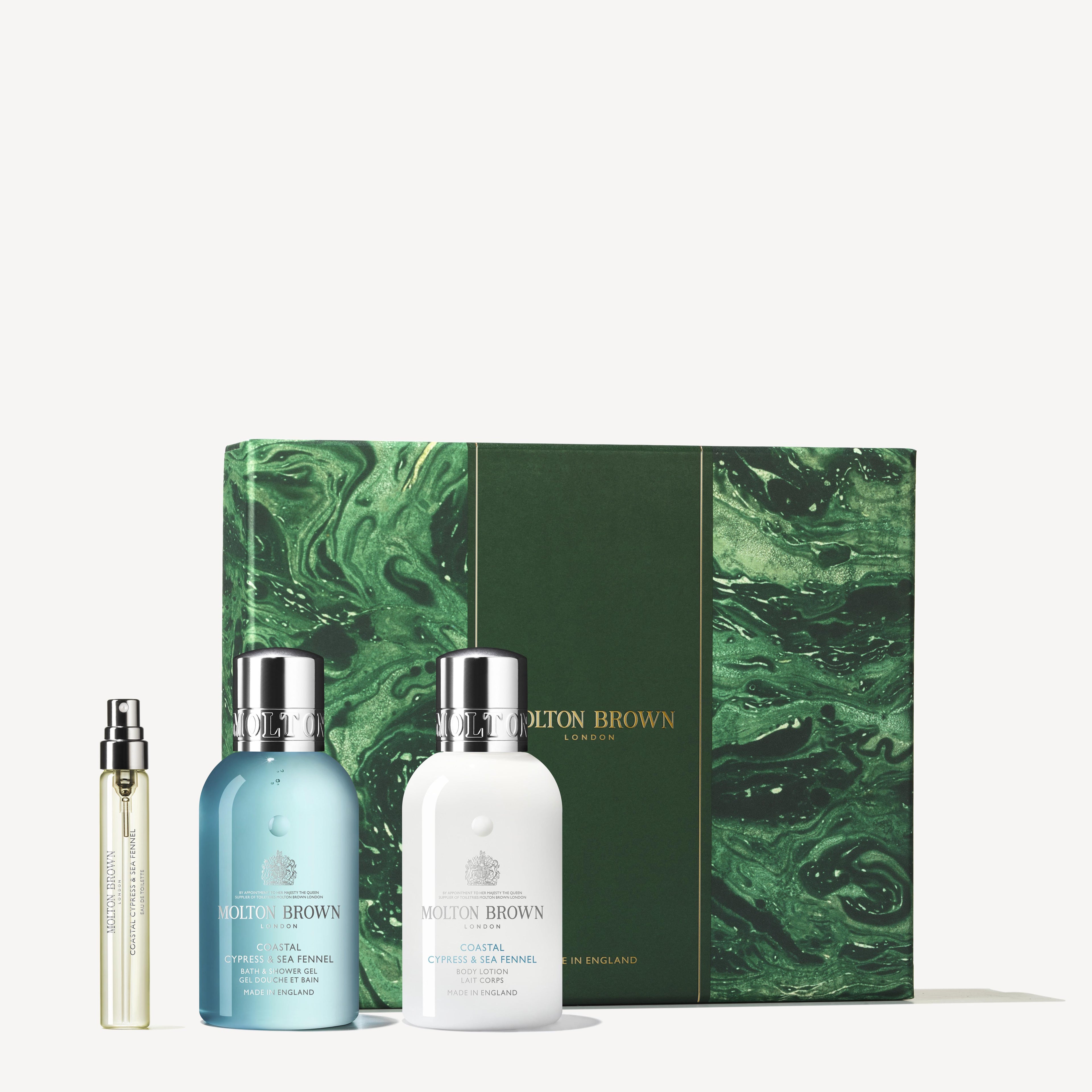 An elegant Coastal Cypress & Sea Fennel Travel Gift Set from MoltonBrown Cyprus, presented in a green and white adorned box with a captivating swirling green marble pattern infused with marine notes. The set includes a small glass vial of Eau de Toilette and two bottles with rounded caps, all labeled in blue and white.